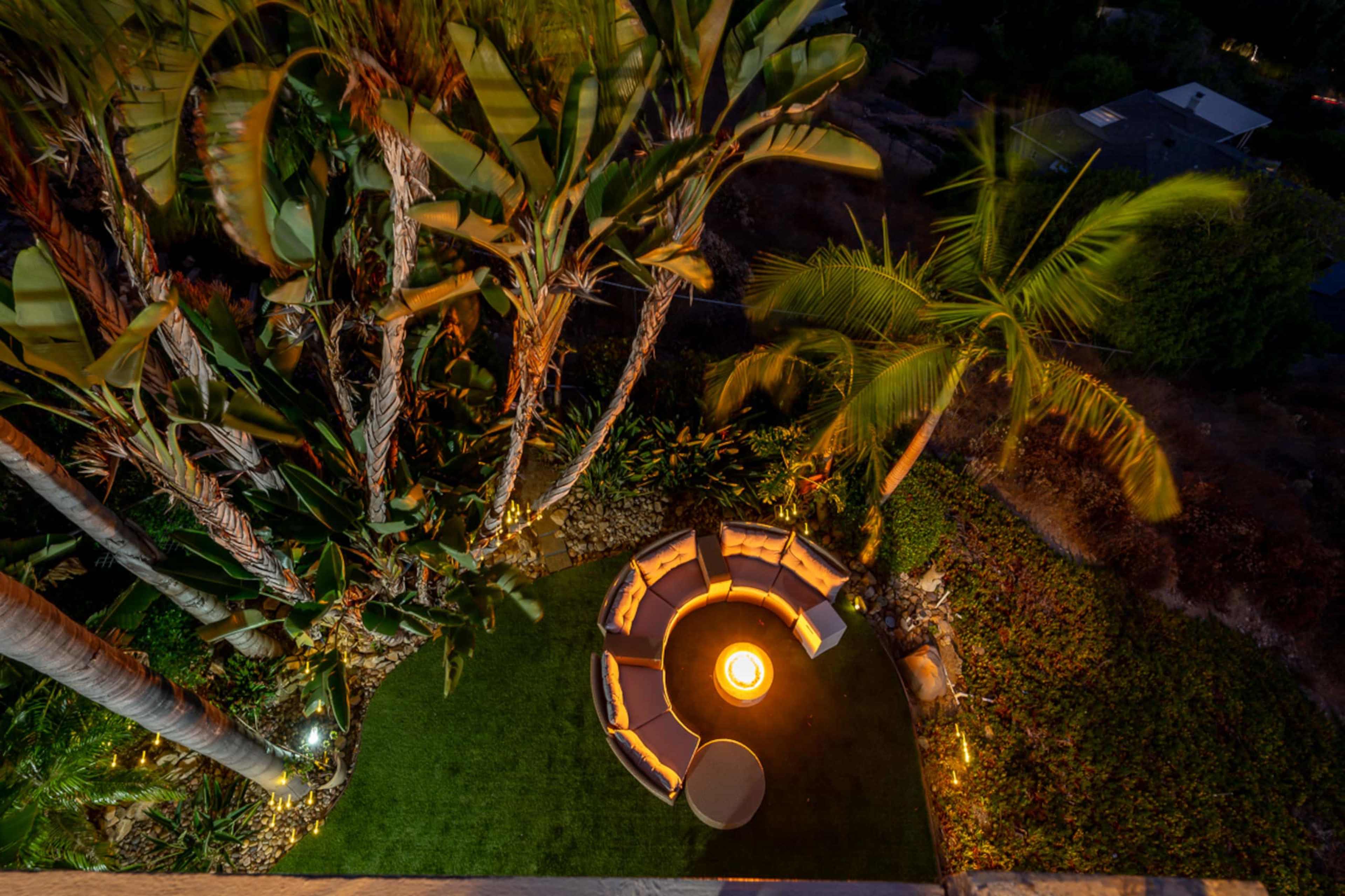 The image depicts a circular outdoor seating area surrounded by tropical plants, featuring a central fire pit illuminated at night.