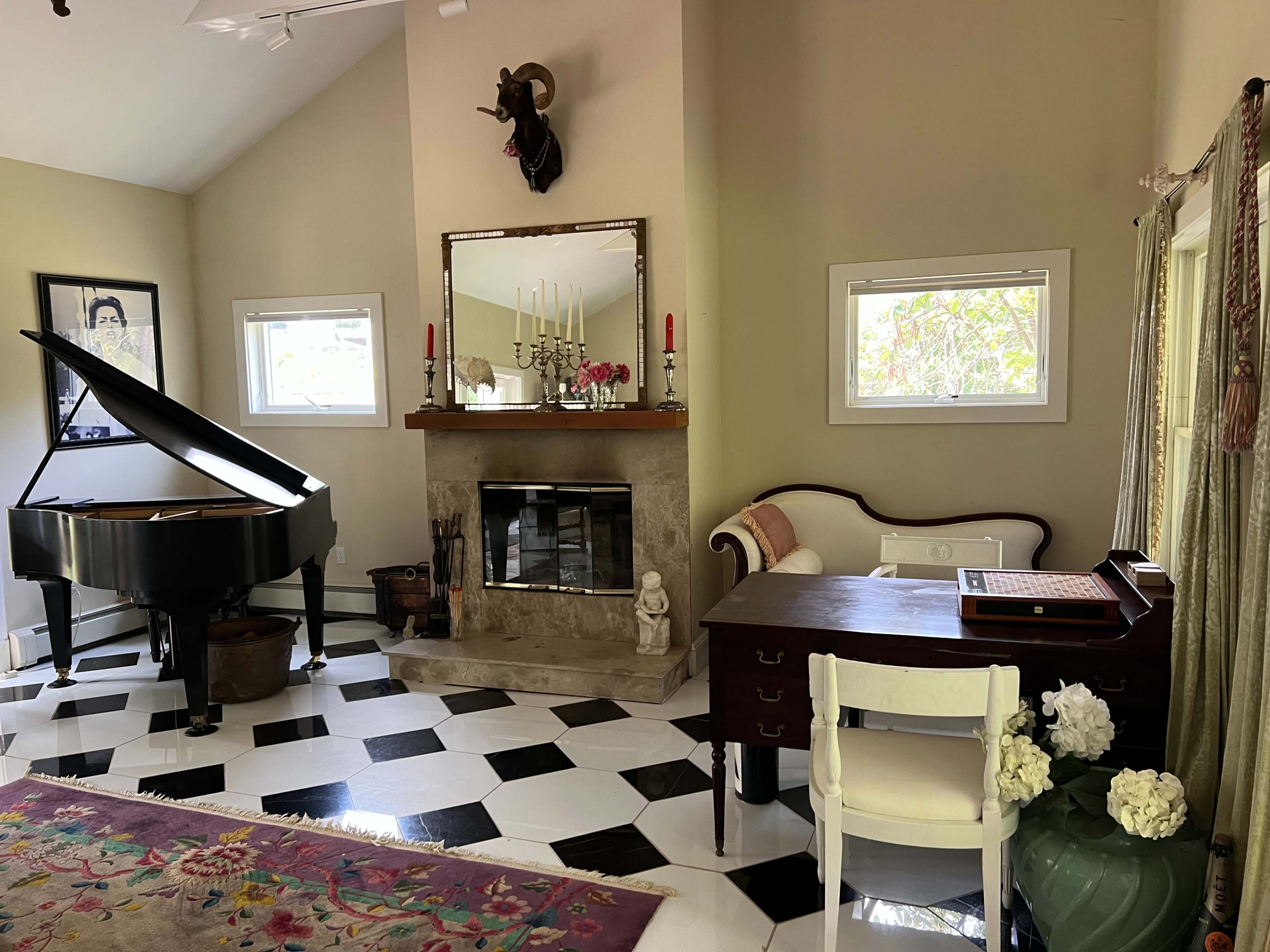 A living room featuring a grand piano, a fireplace with a mounted animal head, a vintage-style sofa, a wooden desk, and a patterned area rug on a checkered floor.