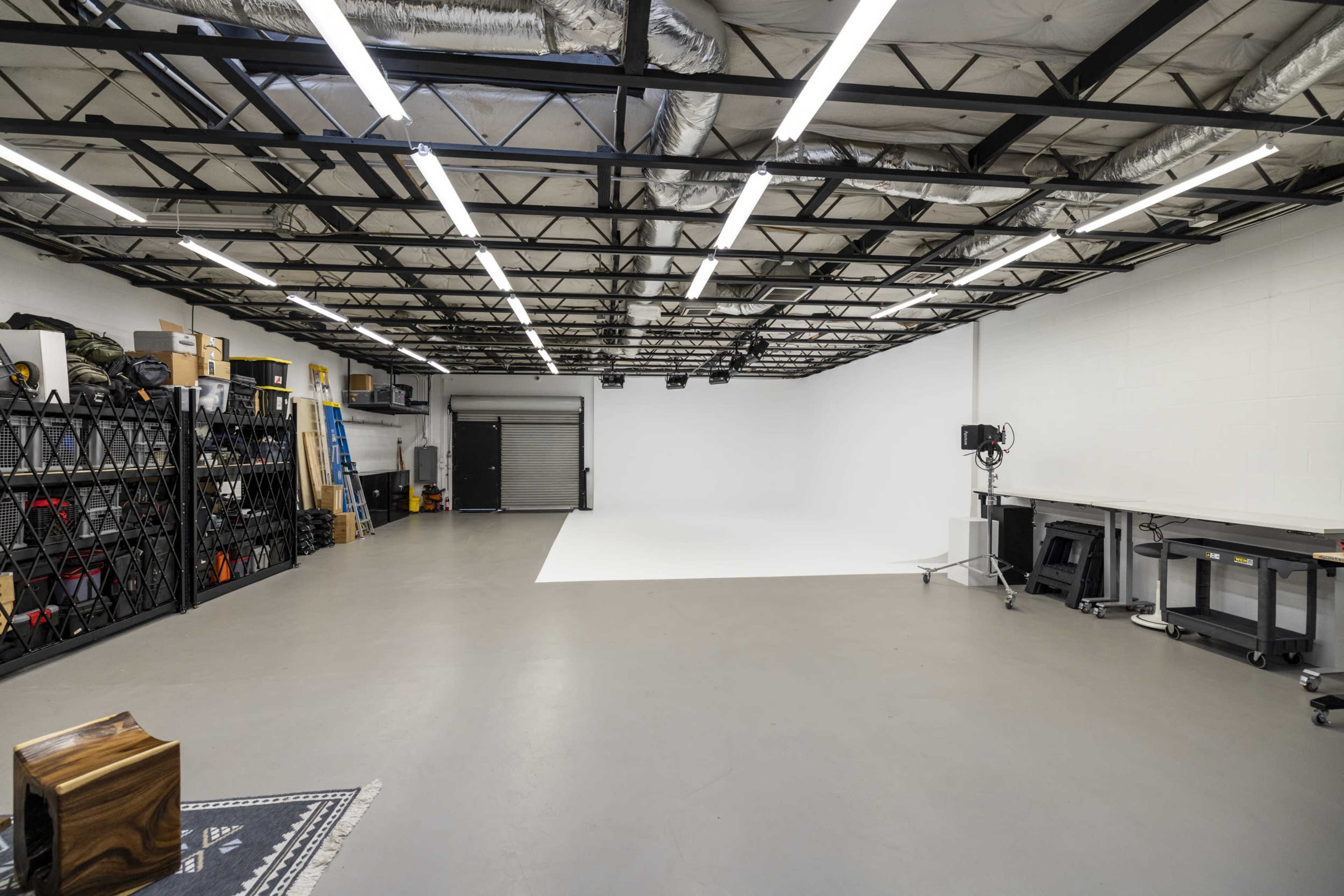 The image shows a spacious, brightly lit studio with a white backdrop, storage shelves, and various equipment neatly organized along the walls.