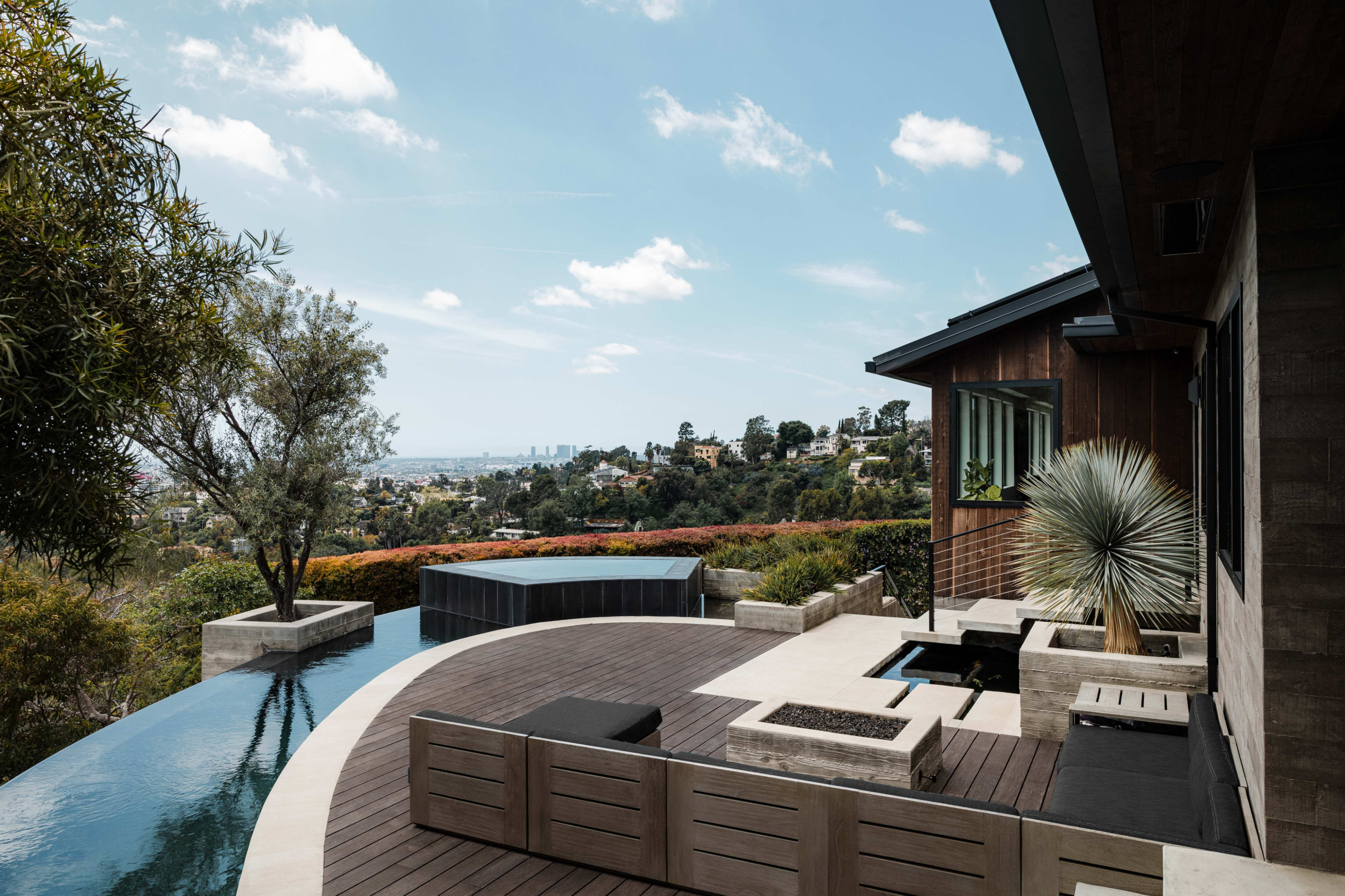 A modern home features an infinity pool, wooden decking, and landscaped gardens with views of the city in the background.