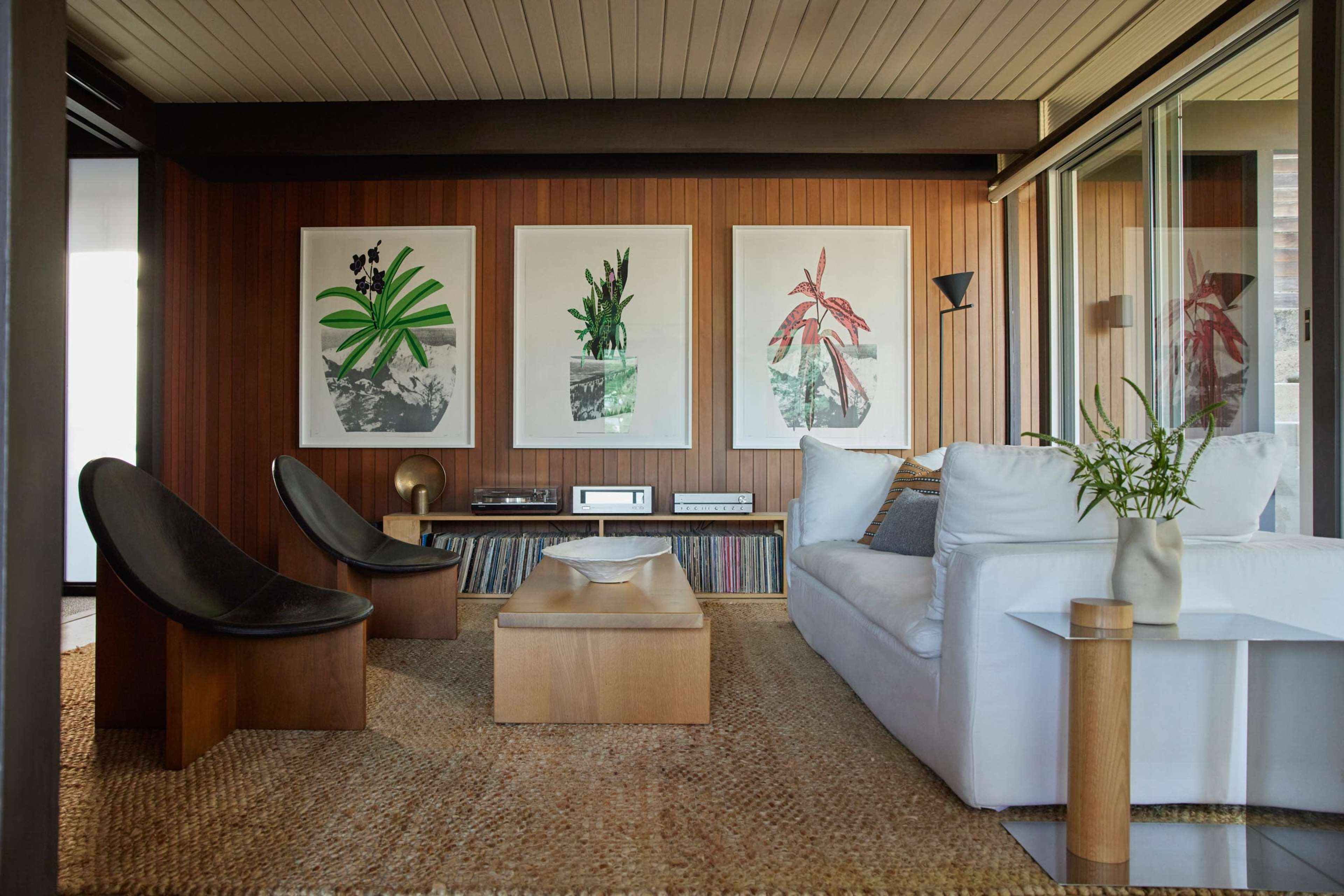 A modern living room features two curved chairs, a white sofa, and three large framed botanical prints on wooden walls, with a coffee table and a record player on display.