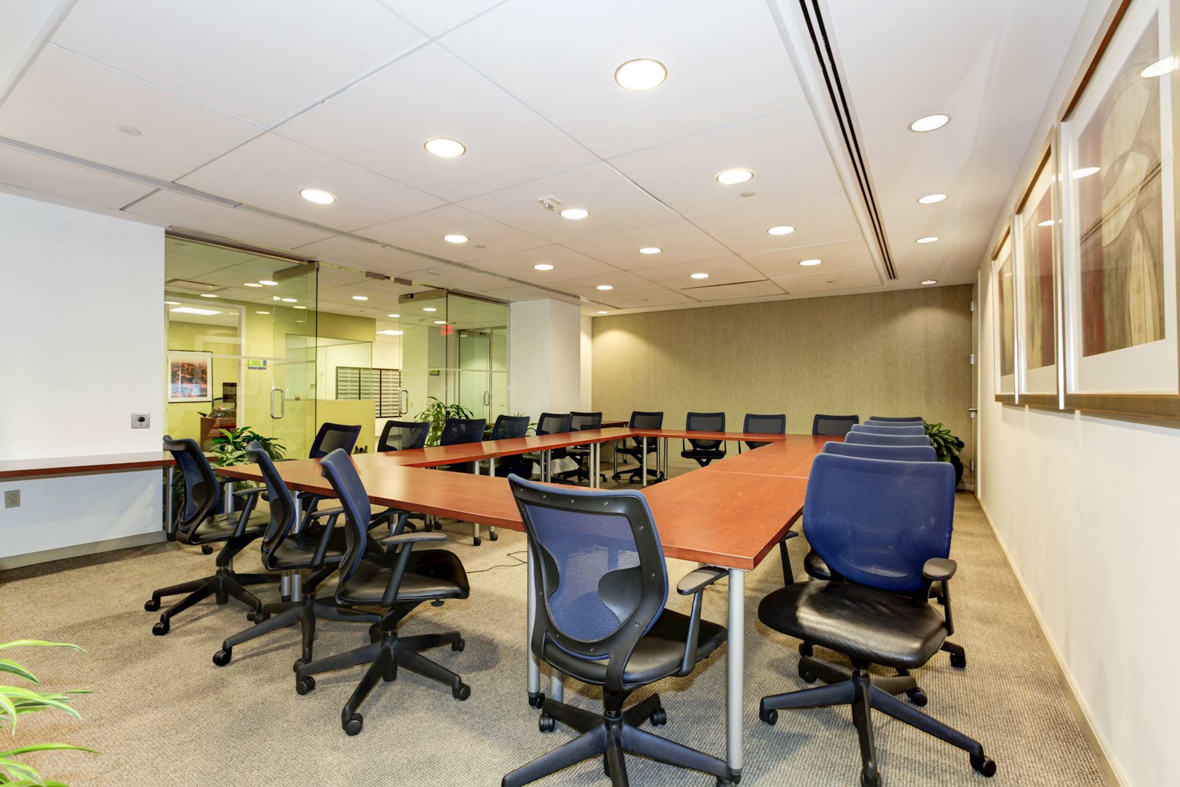 A large conference room features a rectangular table surrounded by office chairs, with glass walls and potted plants providing a view of an adjoining area.