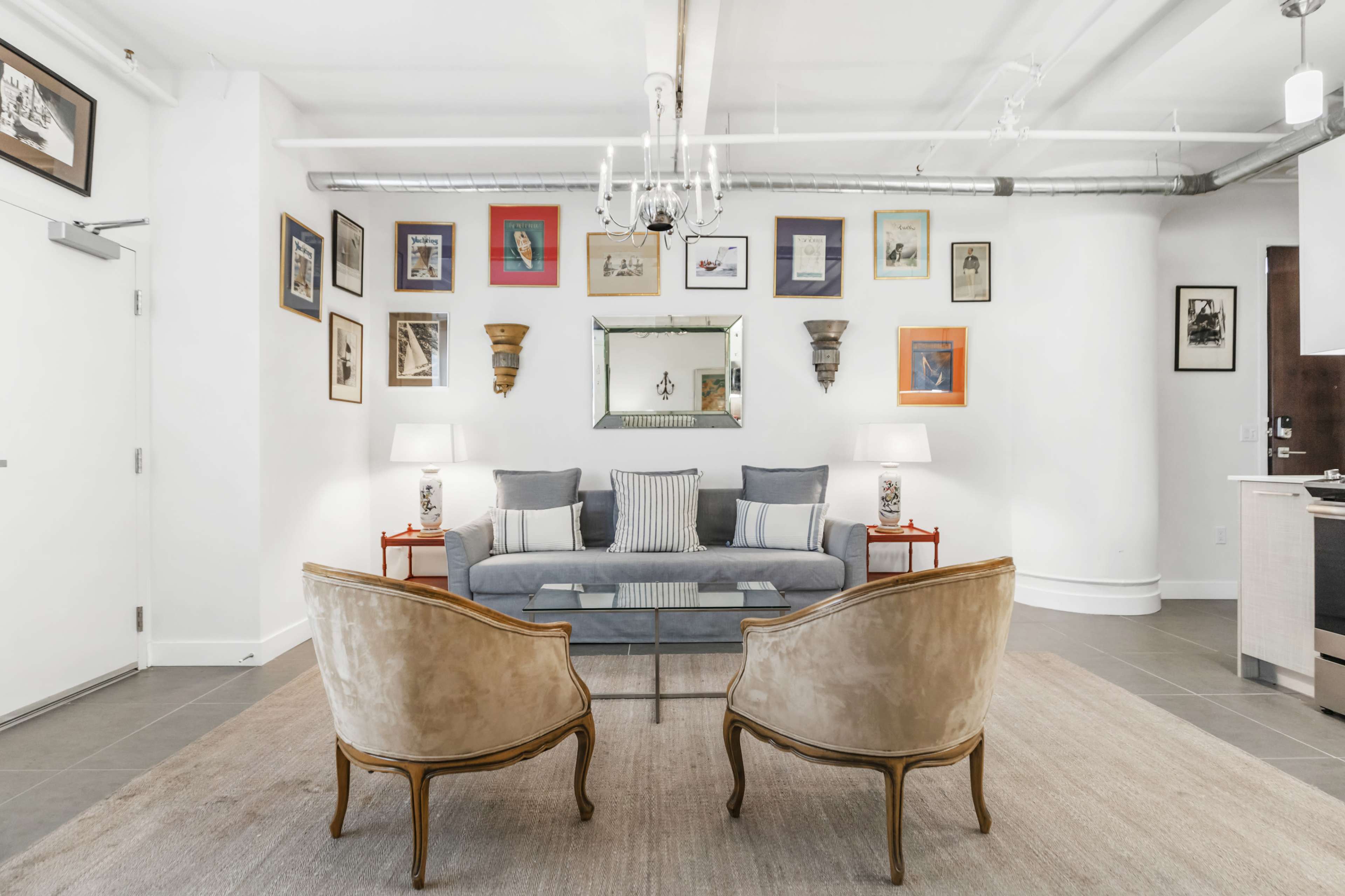 A modern living room features a gray sofa flanked by two elegant chairs, surrounded by framed artwork on the walls and illuminated by lamps on side tables.