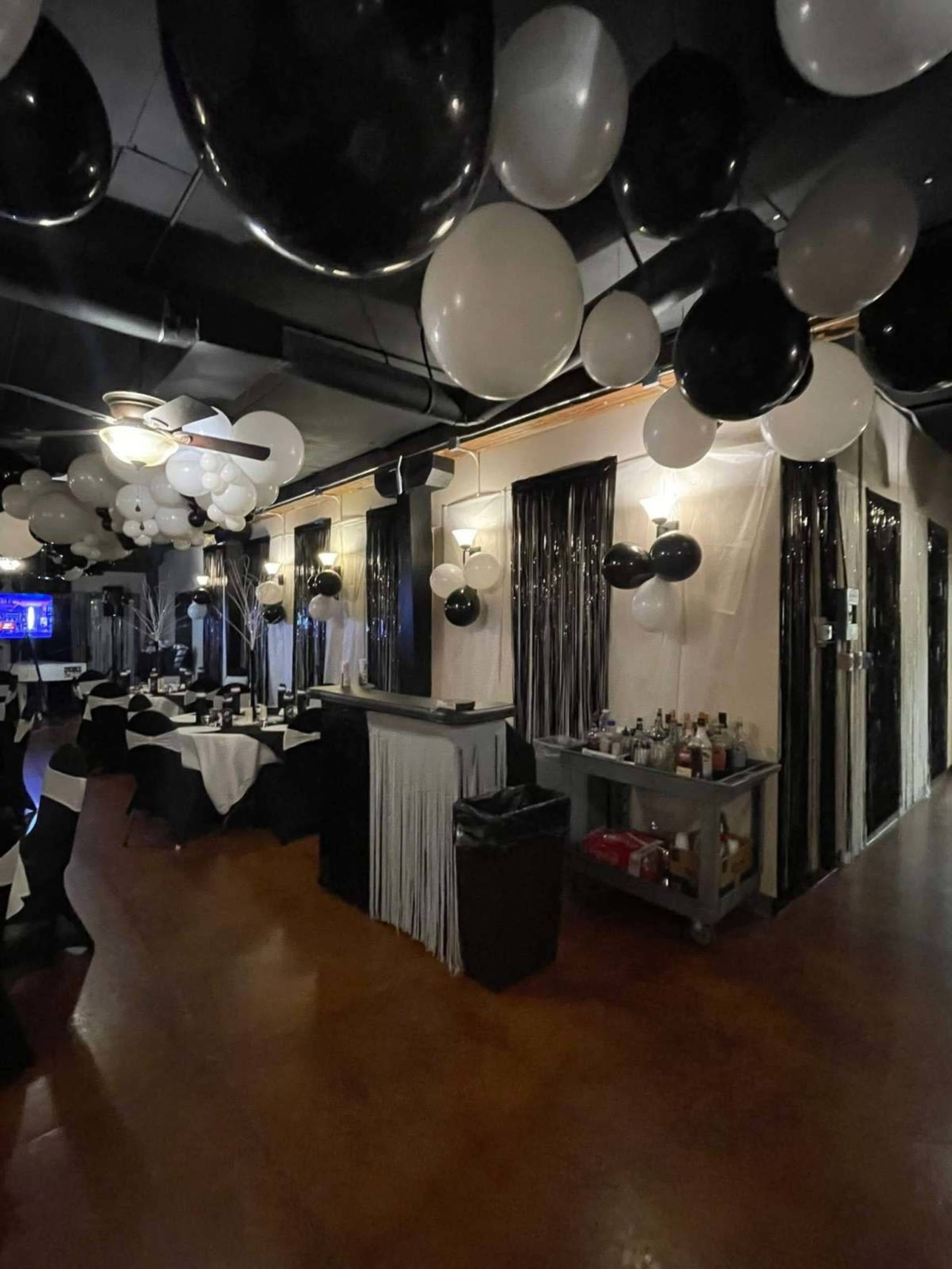 The image shows a decorated restaurant interior with black and white balloons hanging from the ceiling and tables set for dining.