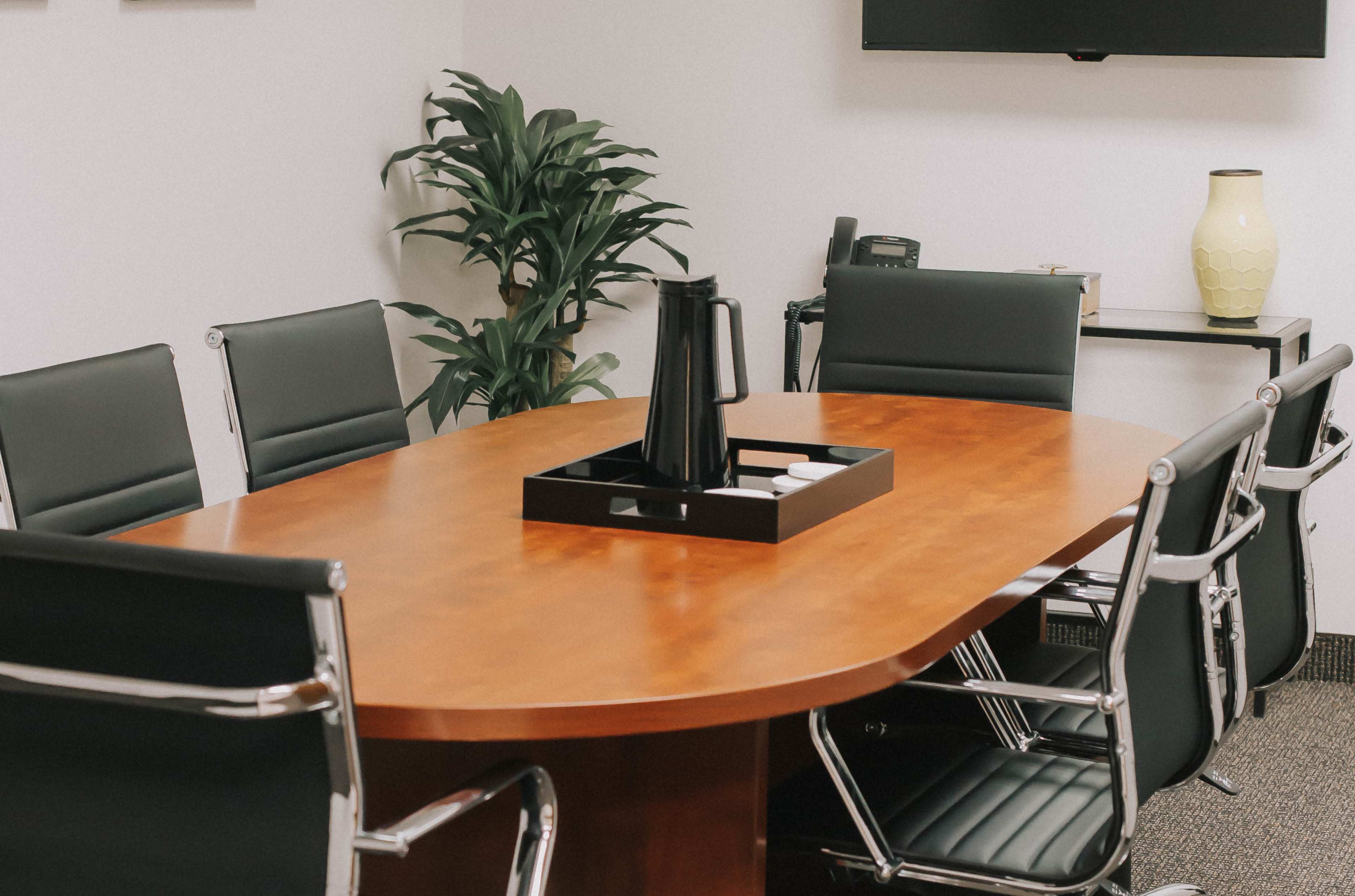 A wooden conference table surrounded by black leather chairs is set up in a modern meeting room with a plant in the corner.