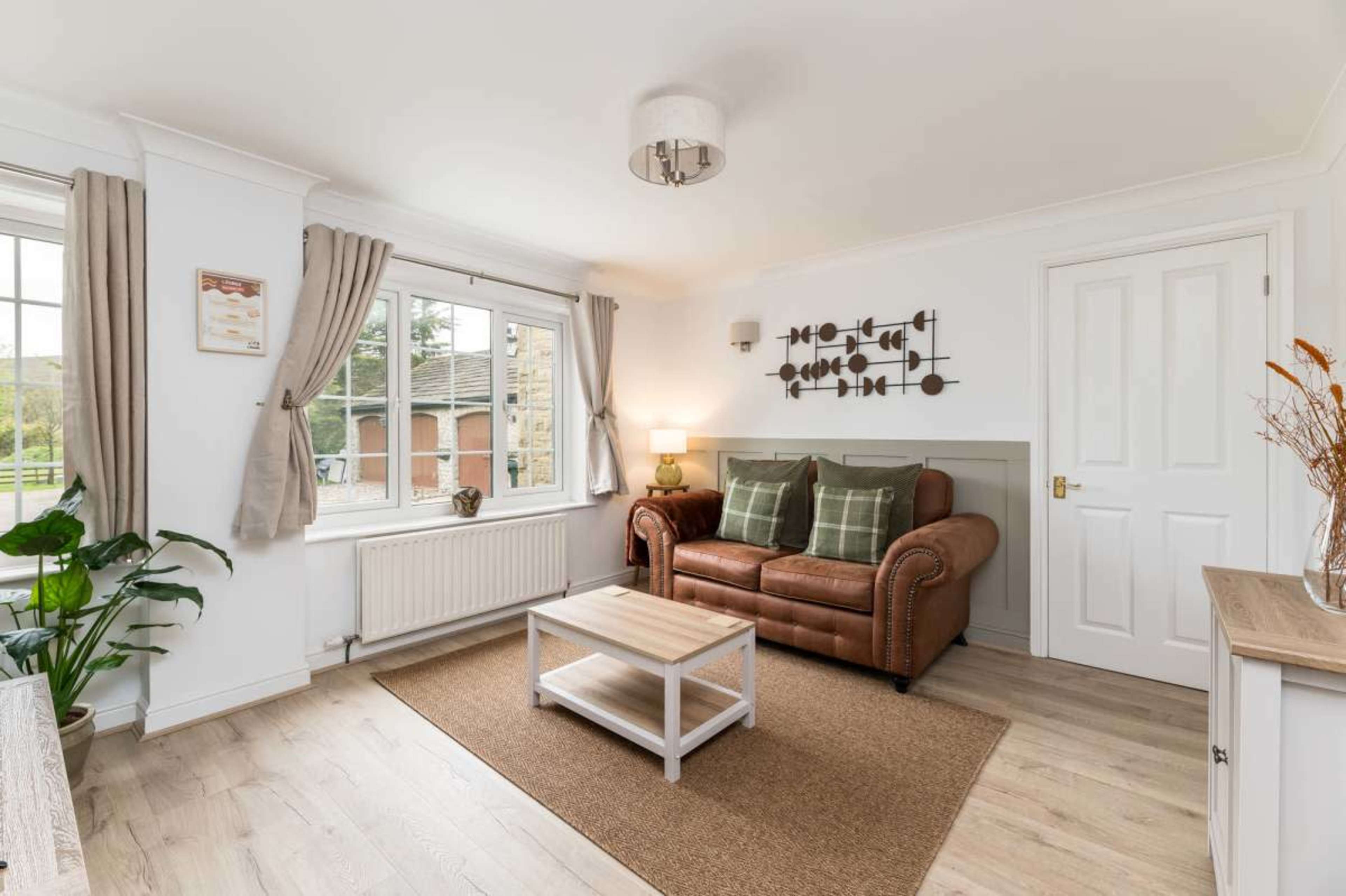 The image shows a cozy living room featuring a brown leather sofa, a small white coffee table, and a large window with beige curtains.