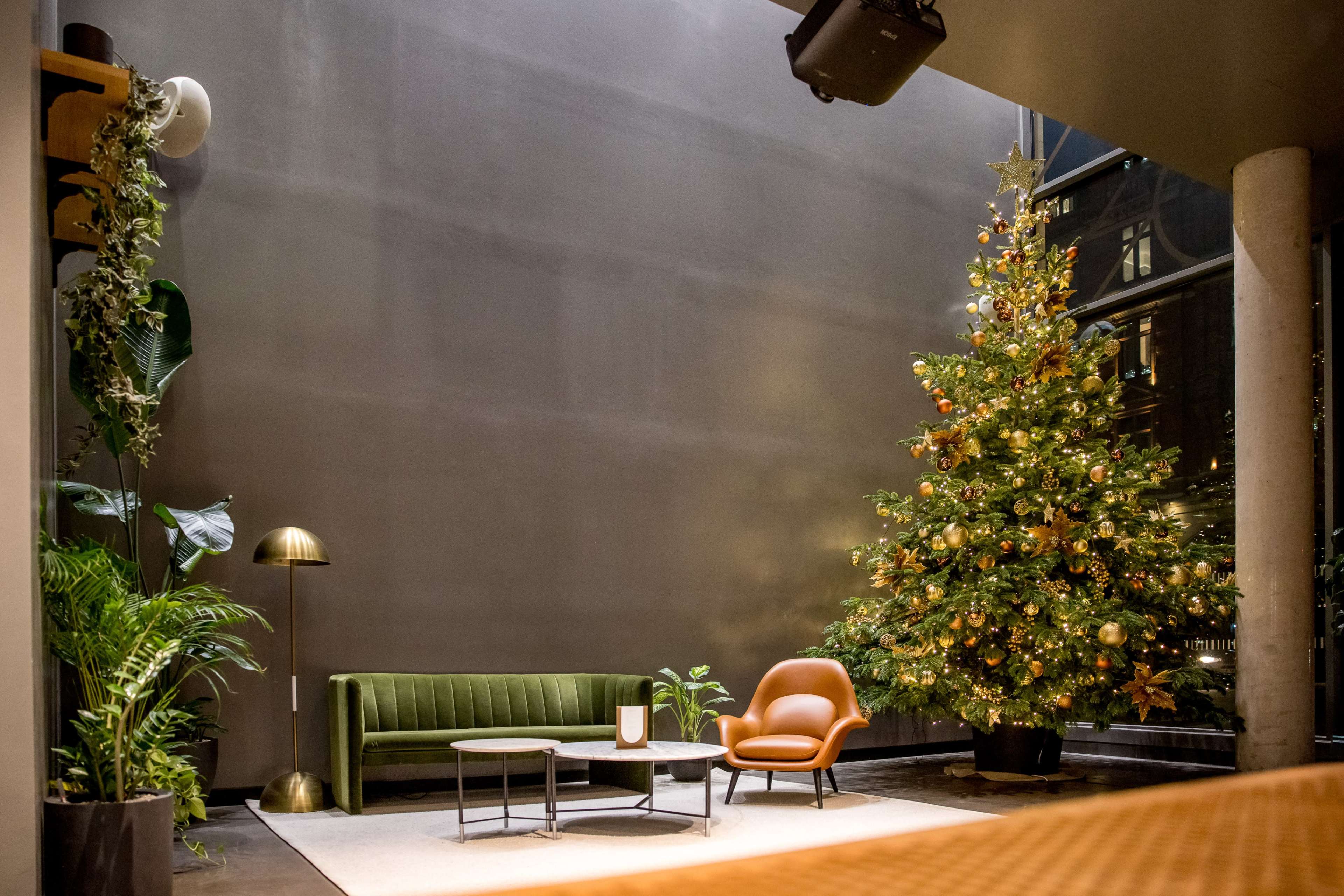 The image shows a modern indoor space featuring a Christmas tree decorated with ornaments, a green sofa, an orange chair, and a standing lamp.