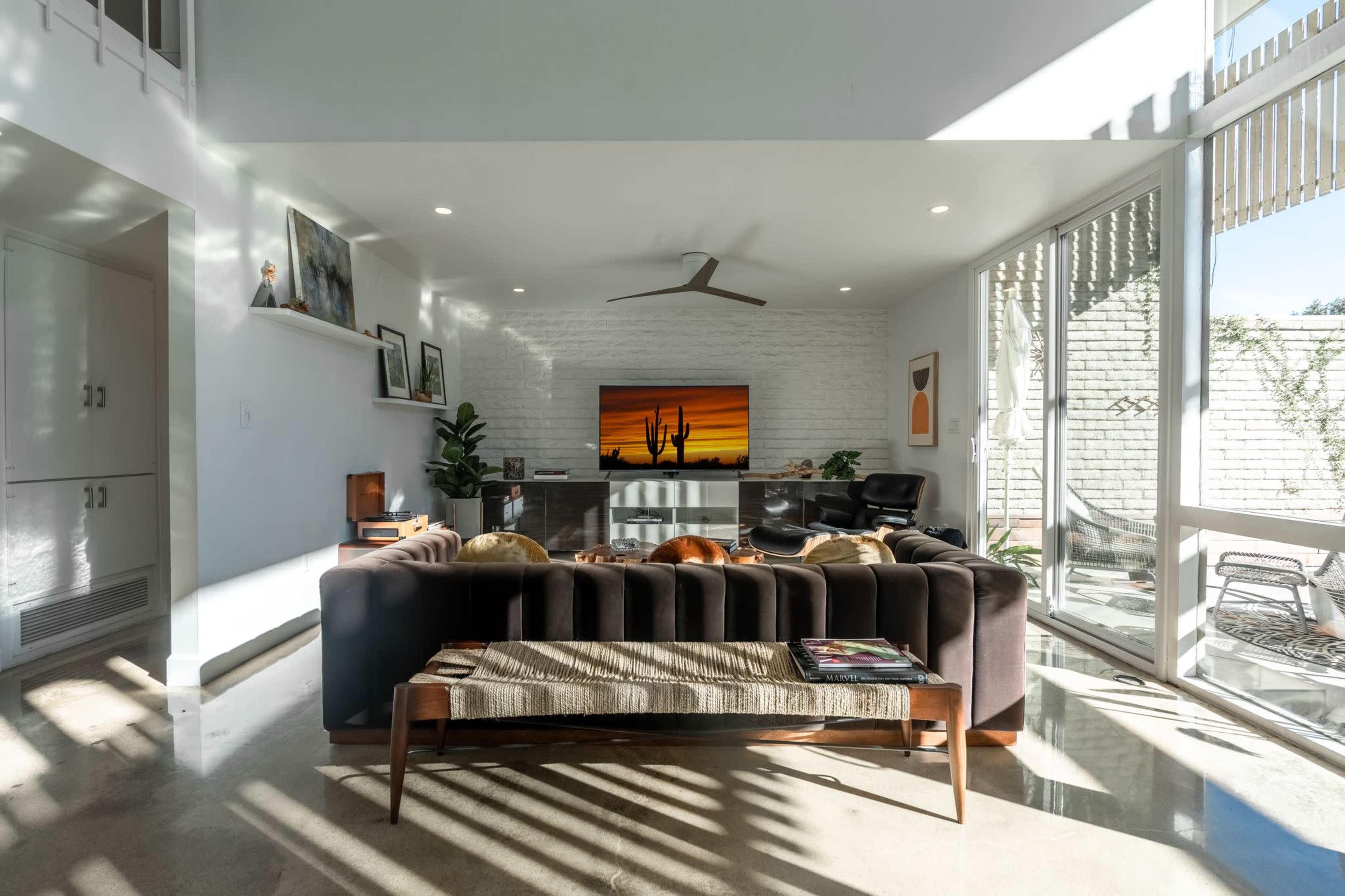 The scene shows a modern living room with a dark couch, a large television displaying a sunset, and large windows allowing natural light to fill the space.