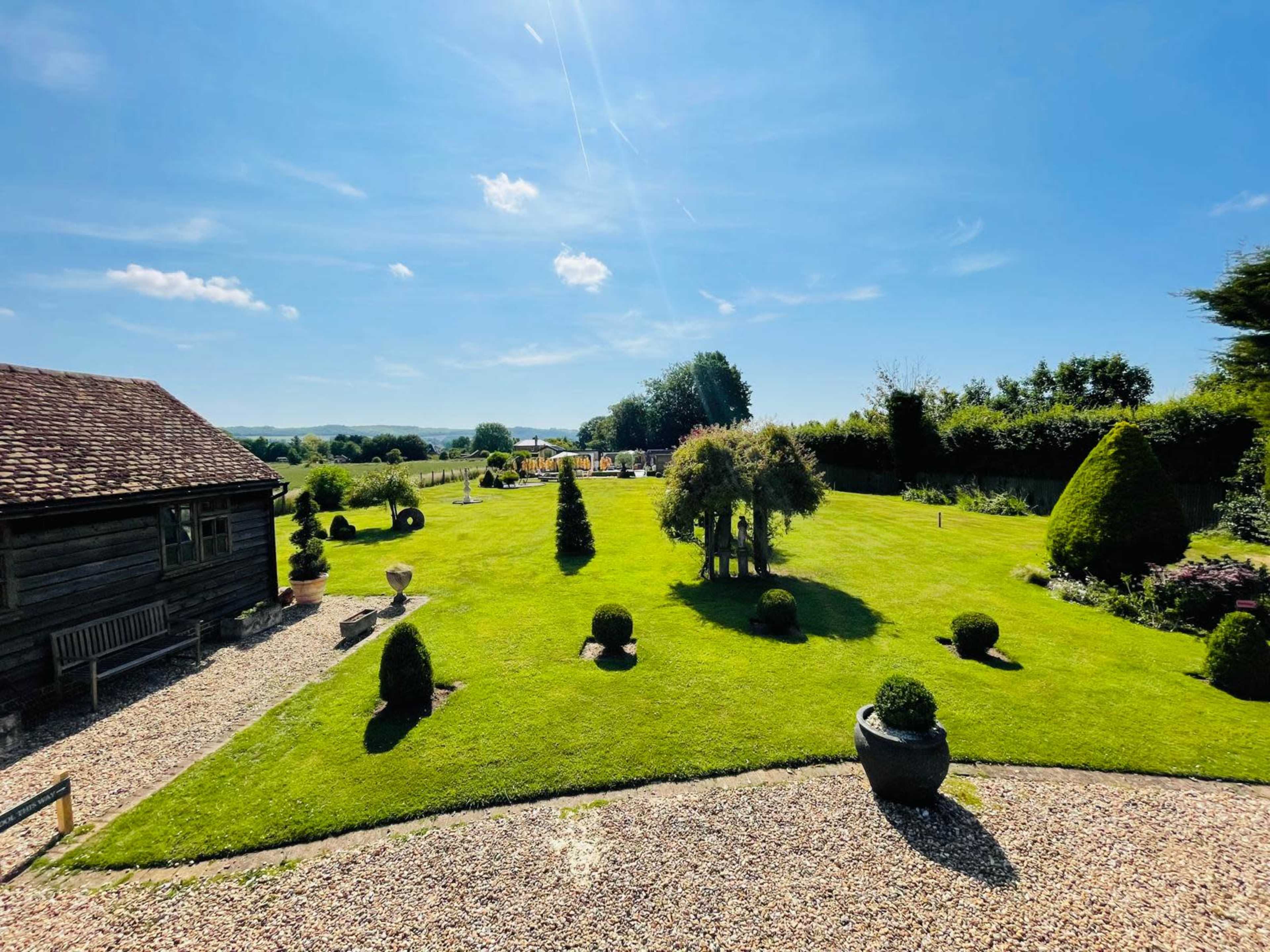 A well-maintained garden features a variety of shrubs and trees, with a gravel path leading to a rustic wooden building under a blue sky.