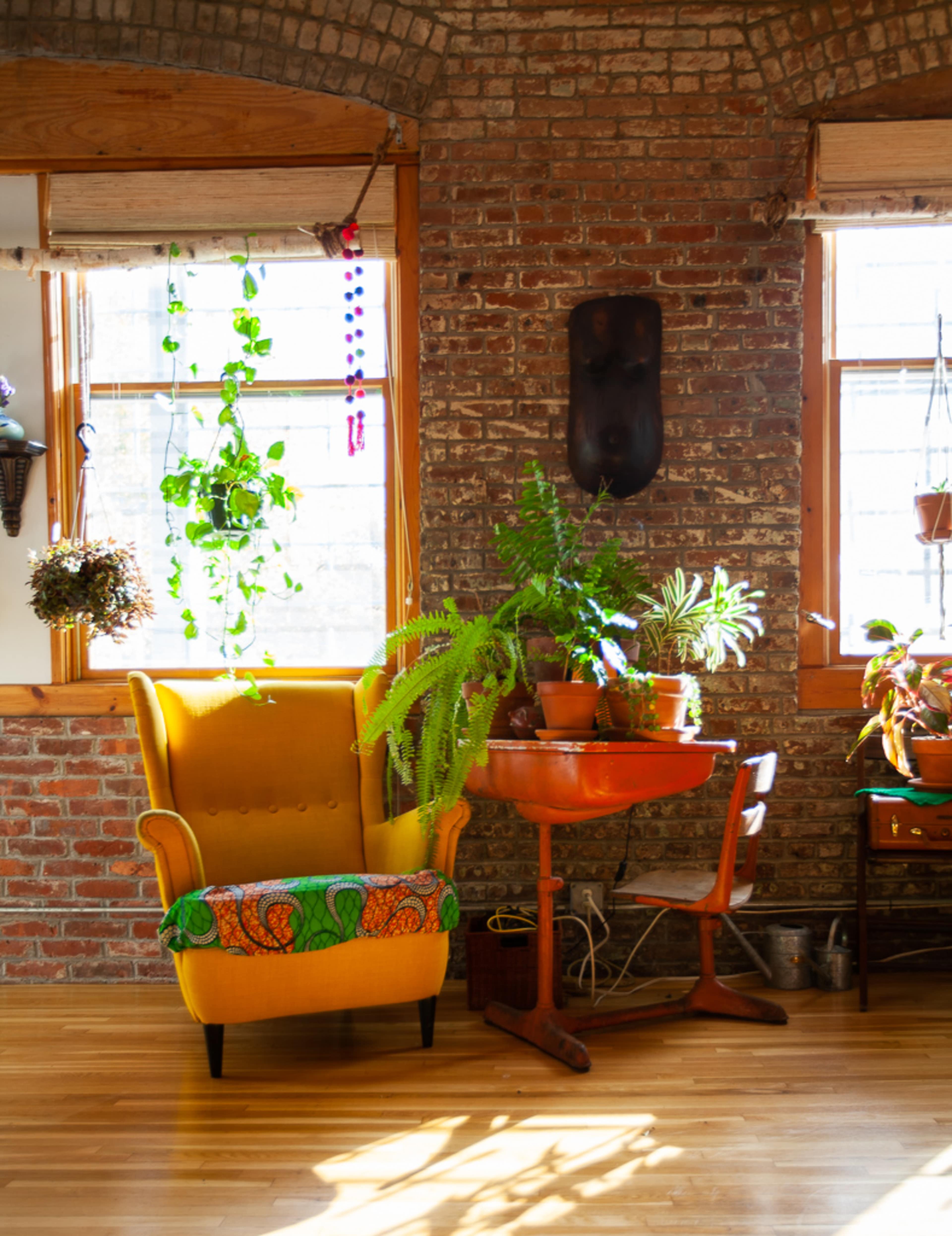 A bright yellow armchair sits beside a wooden table surrounded by various potted plants against a brick wall with large windows.