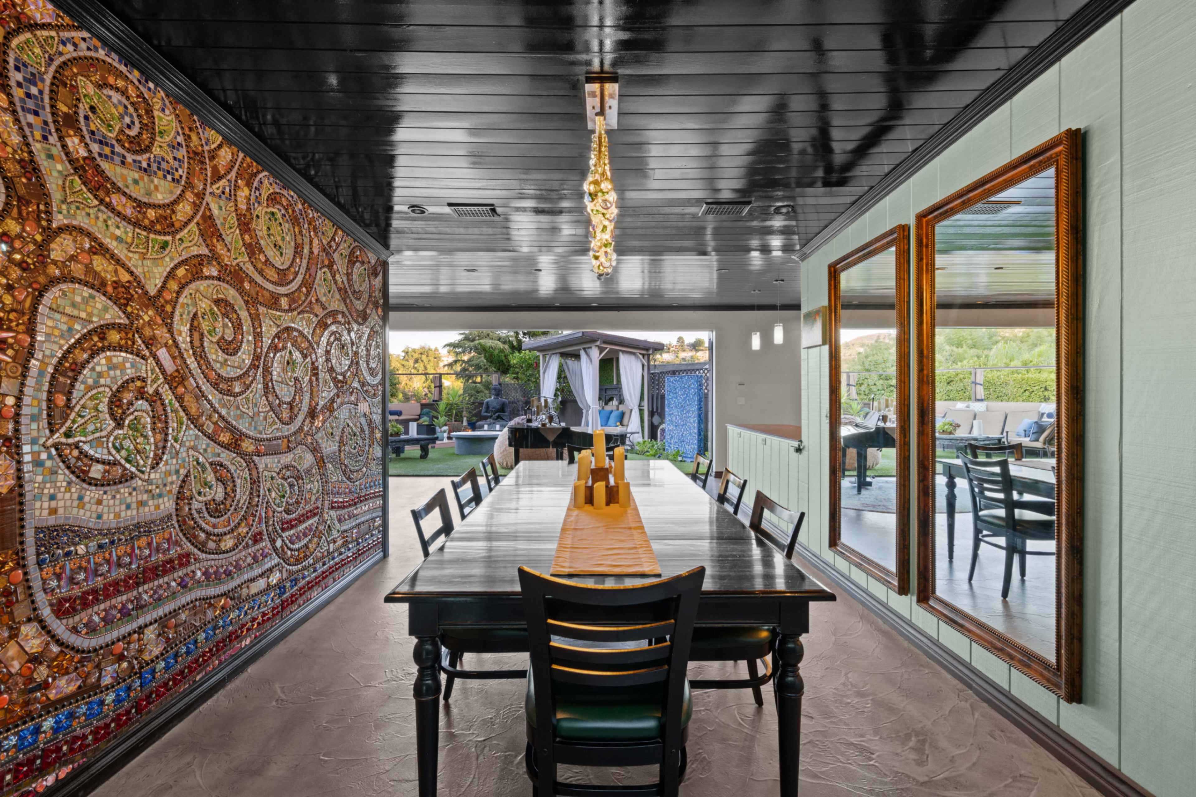 A dining area with a long dark table, surrounded by chairs, a decorative mosaic wall, and a view of an outdoor seating area through large windows.