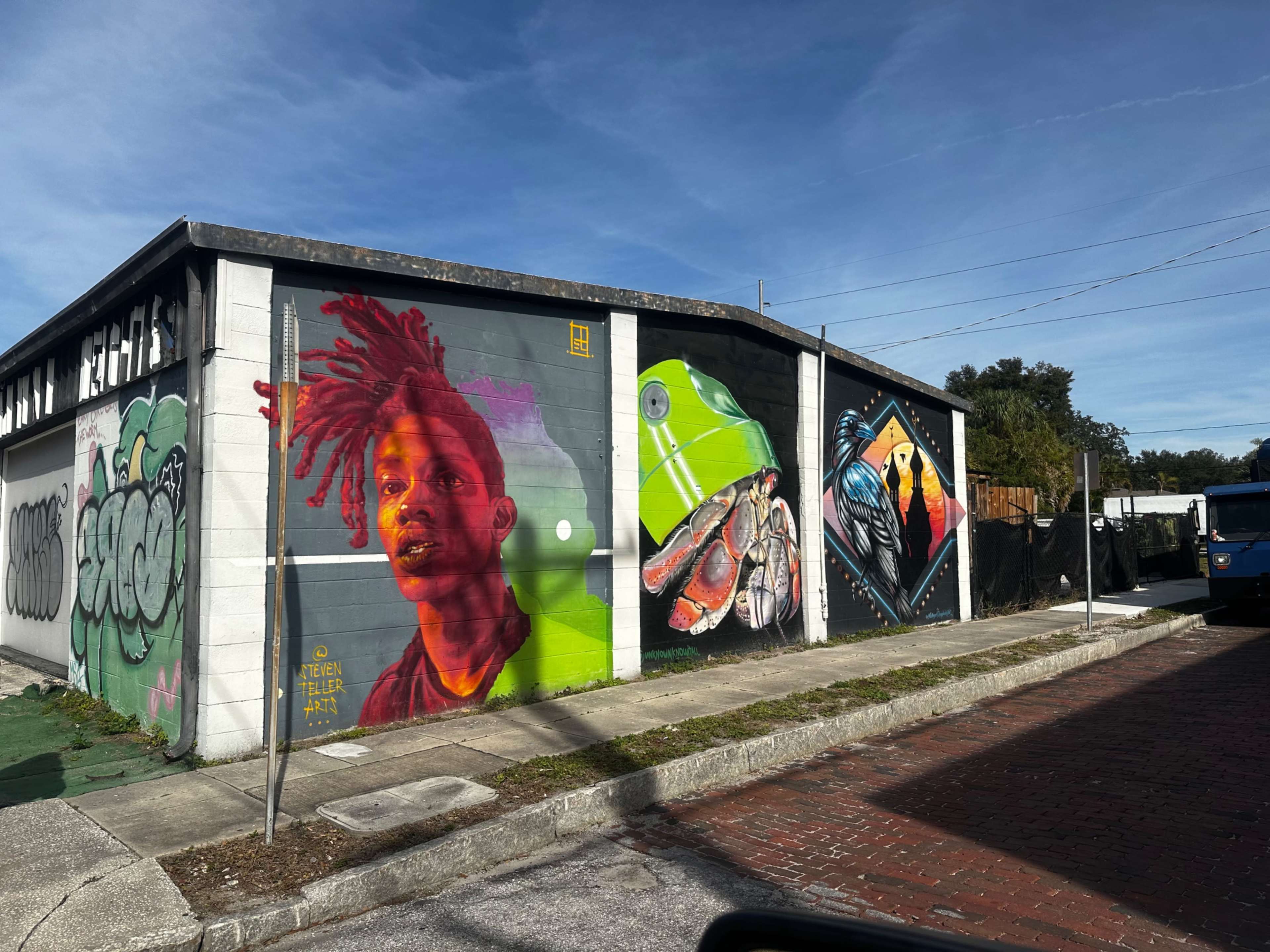 A mural on a building featuring a colorful portrait of a young man alongside various artistic designs, including a hand and abstract shapes.