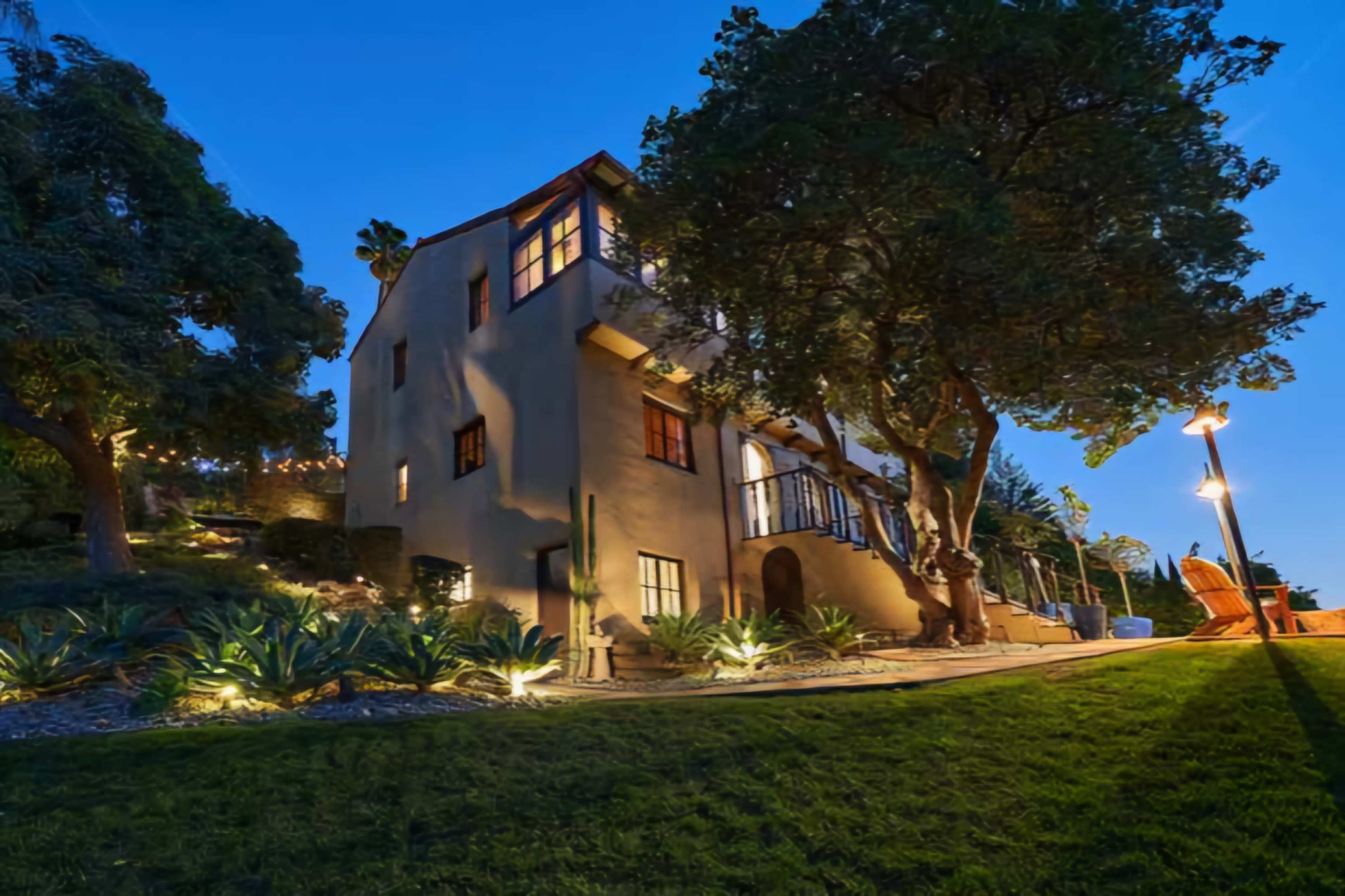 A multi-story house with large windows is set against a twilight sky, surrounded by trees and landscaped greenery.