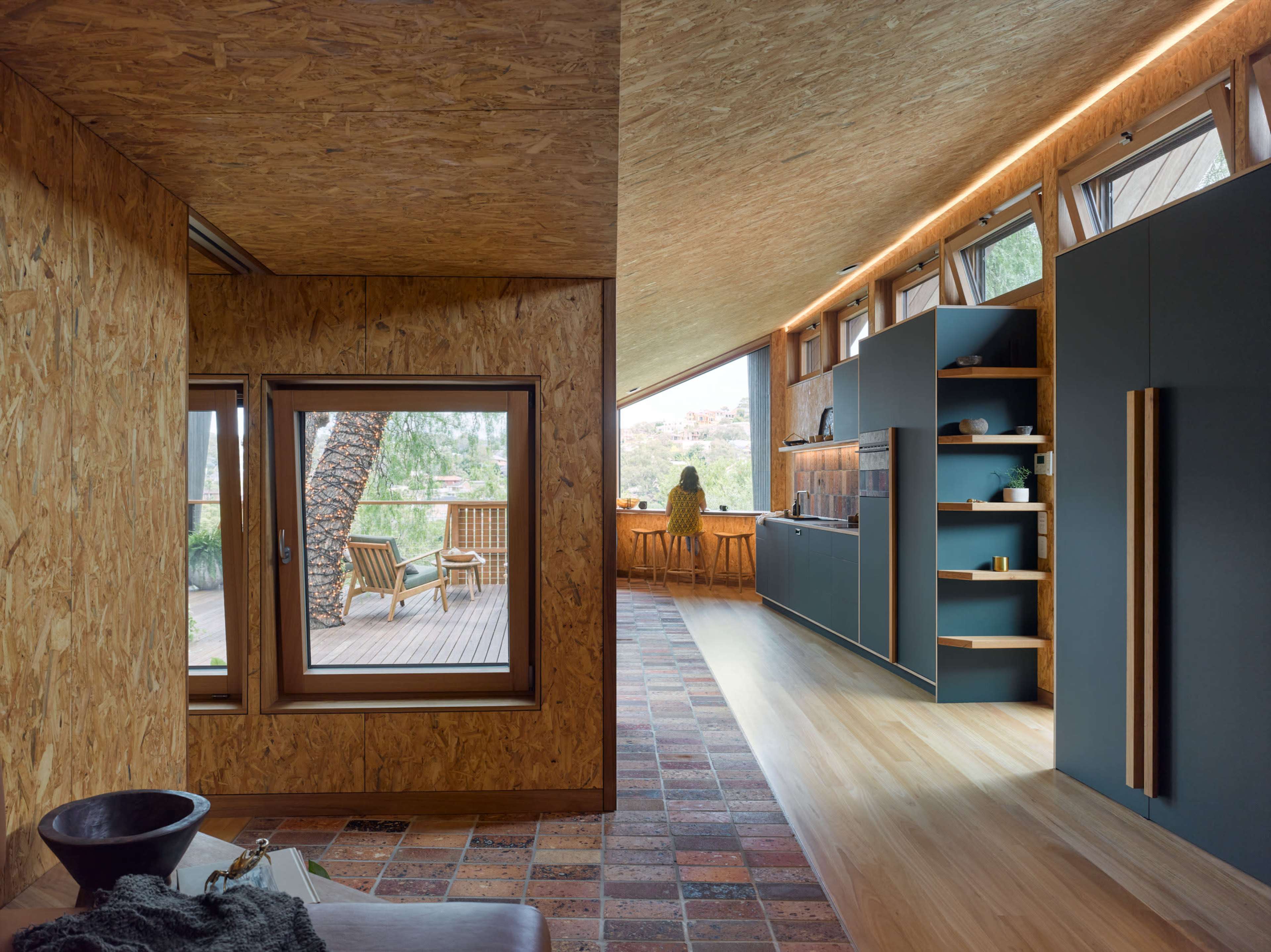 The image shows the interior of a modern home, featuring an open layout with an emphasis on natural materials and a large window that provides a view of the outdoor landscape.