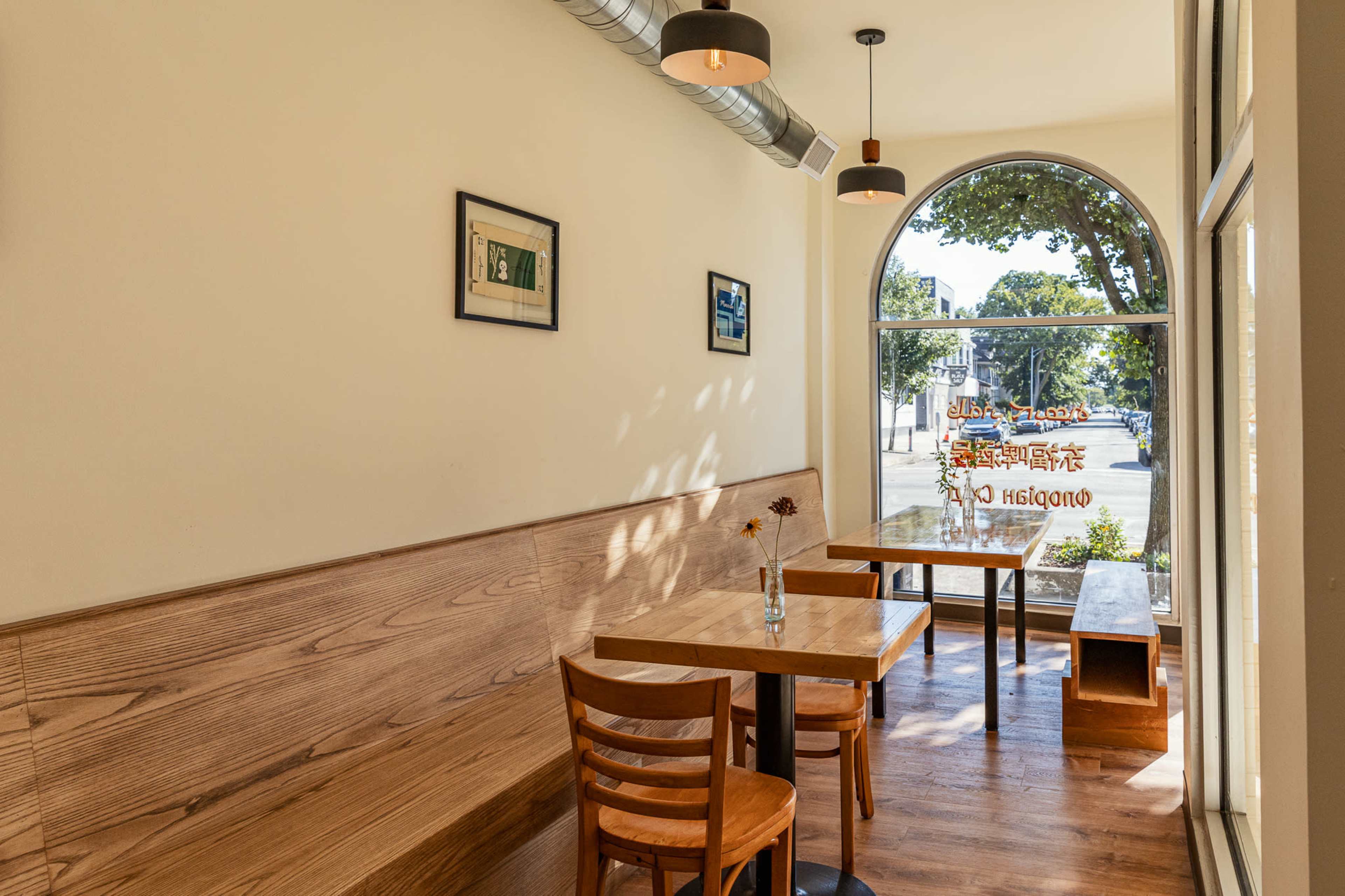 The image shows a simple café interior with wooden seating, tables, and a large arched window that provides natural light.