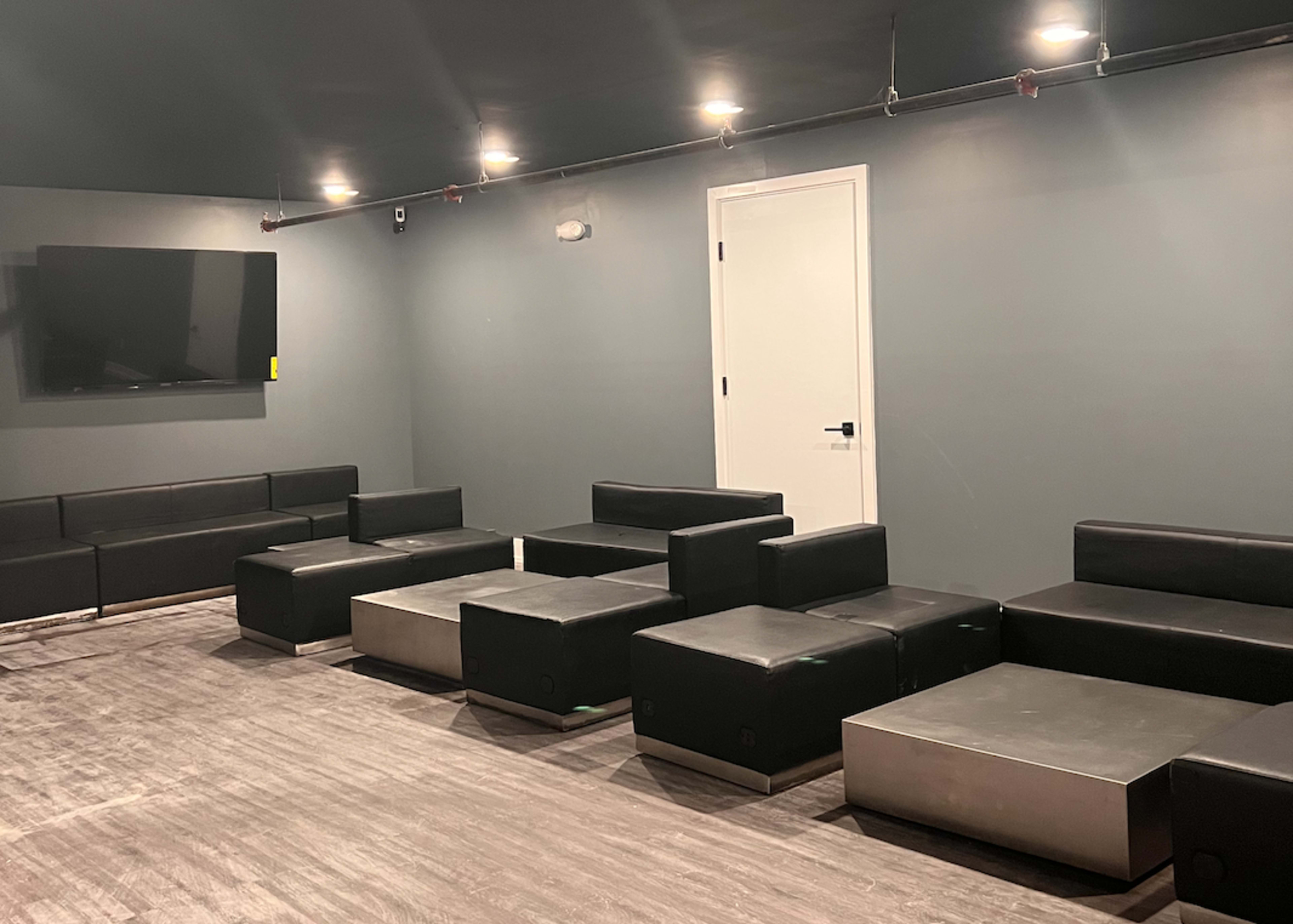 A minimalist lounge area features several black sofas and low tables arranged around a wall-mounted television and a white door.