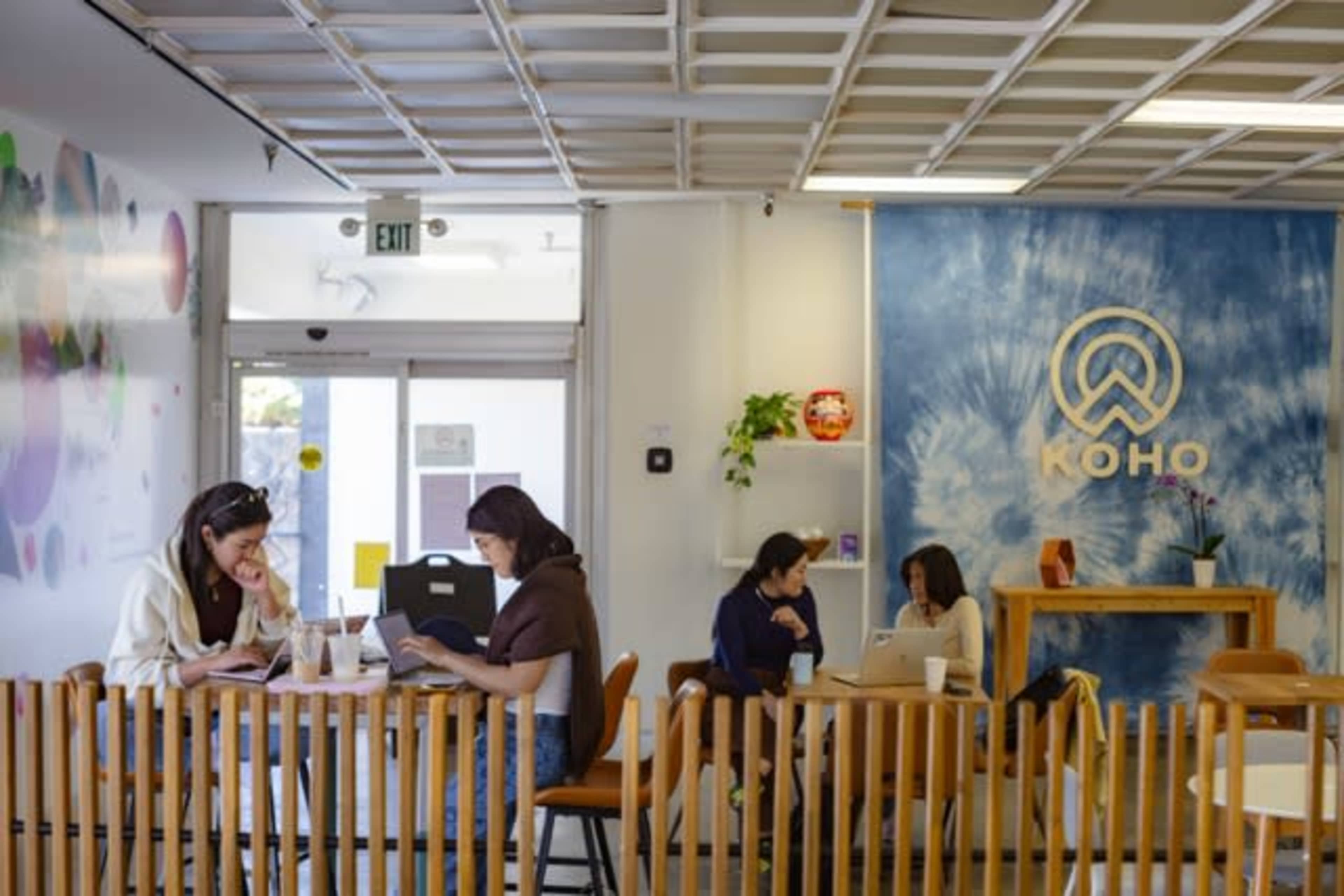The image shows a modern café interior with patrons sitting at tables, some using laptops, and a colorful wall mural in the background.