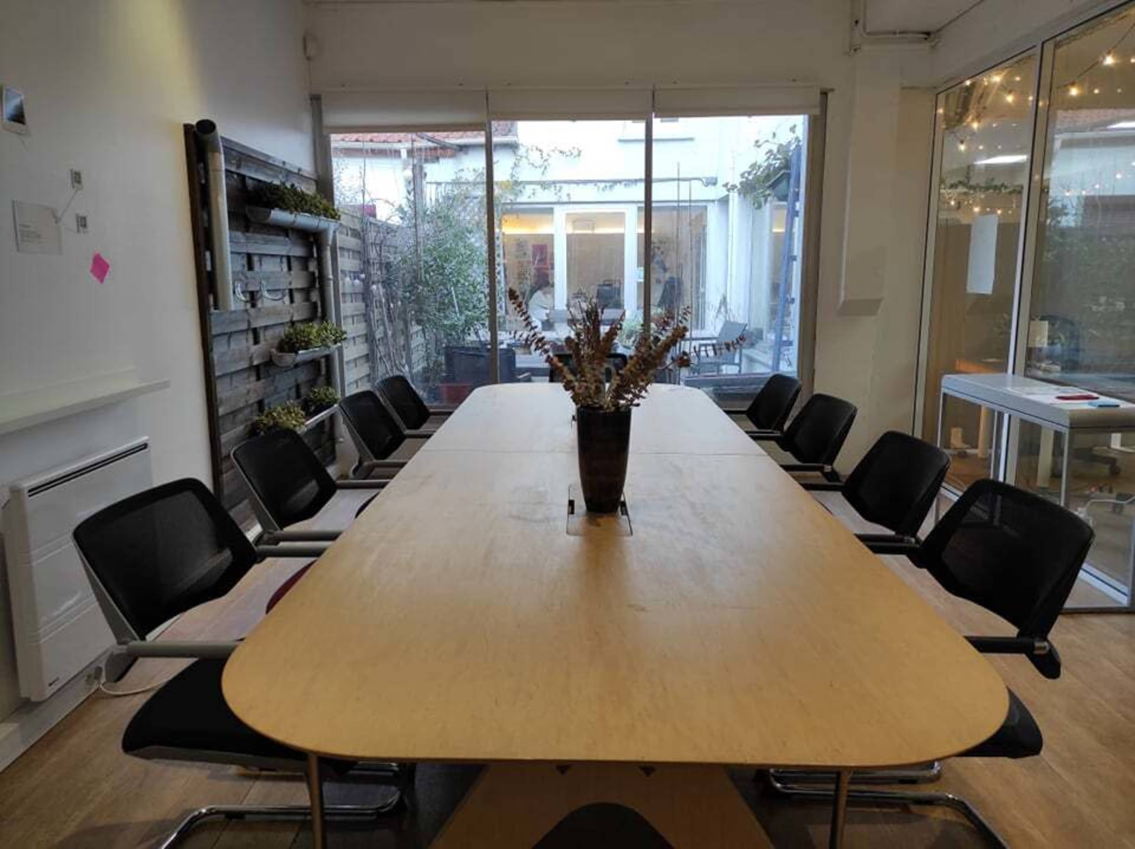 A large wooden conference table is set in a well-lit room with chairs around it, overlooking an outdoor patio through glass walls.