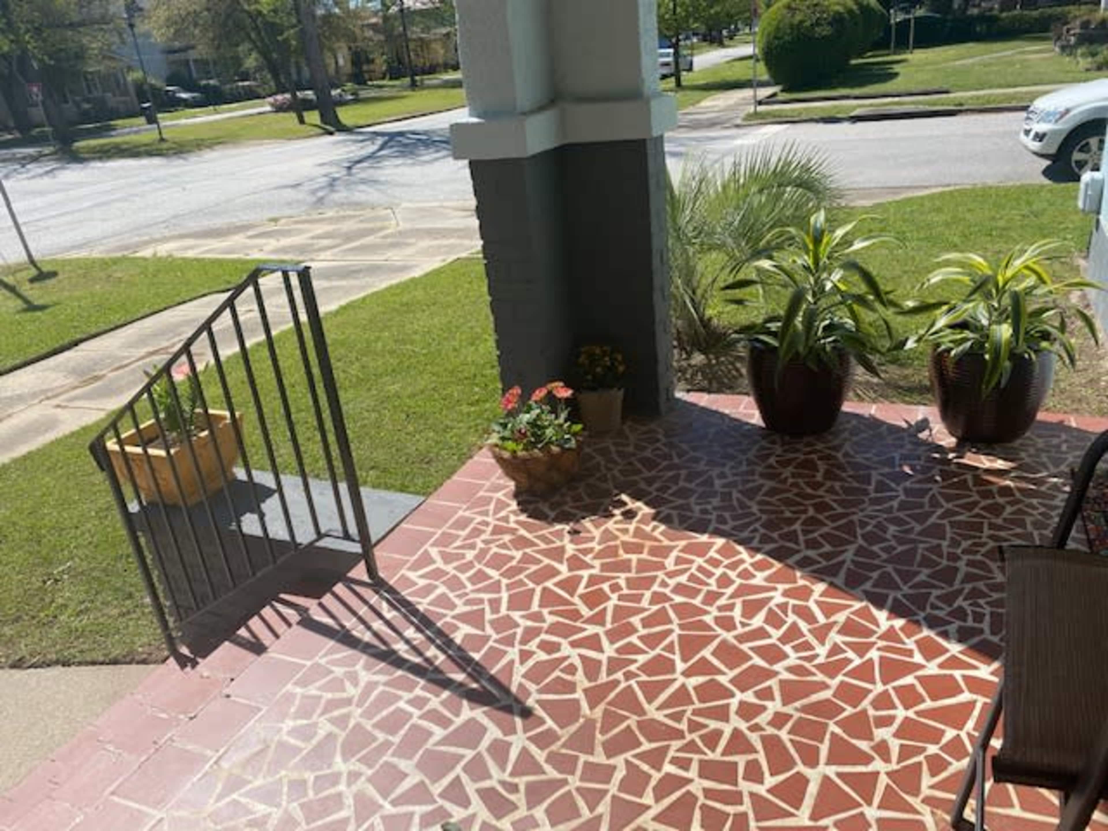 The image shows a porch with a patterned tile floor, potted plants, and a rail leading to the lawn and street.