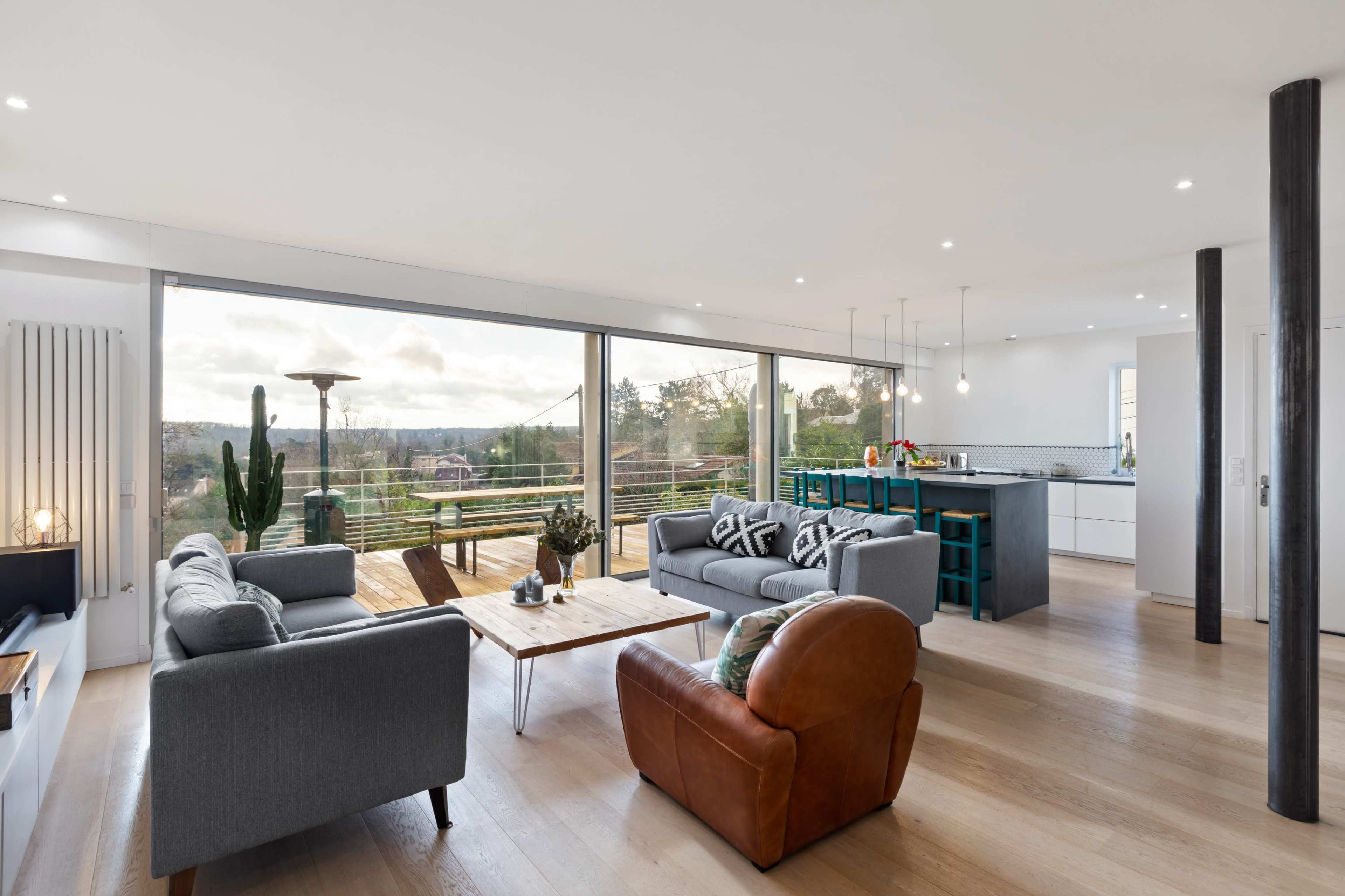 The image shows a modern living room with two gray sofas, an armchair, a coffee table, and a kitchen visible in the background, featuring large windows that overlook a scenic view.