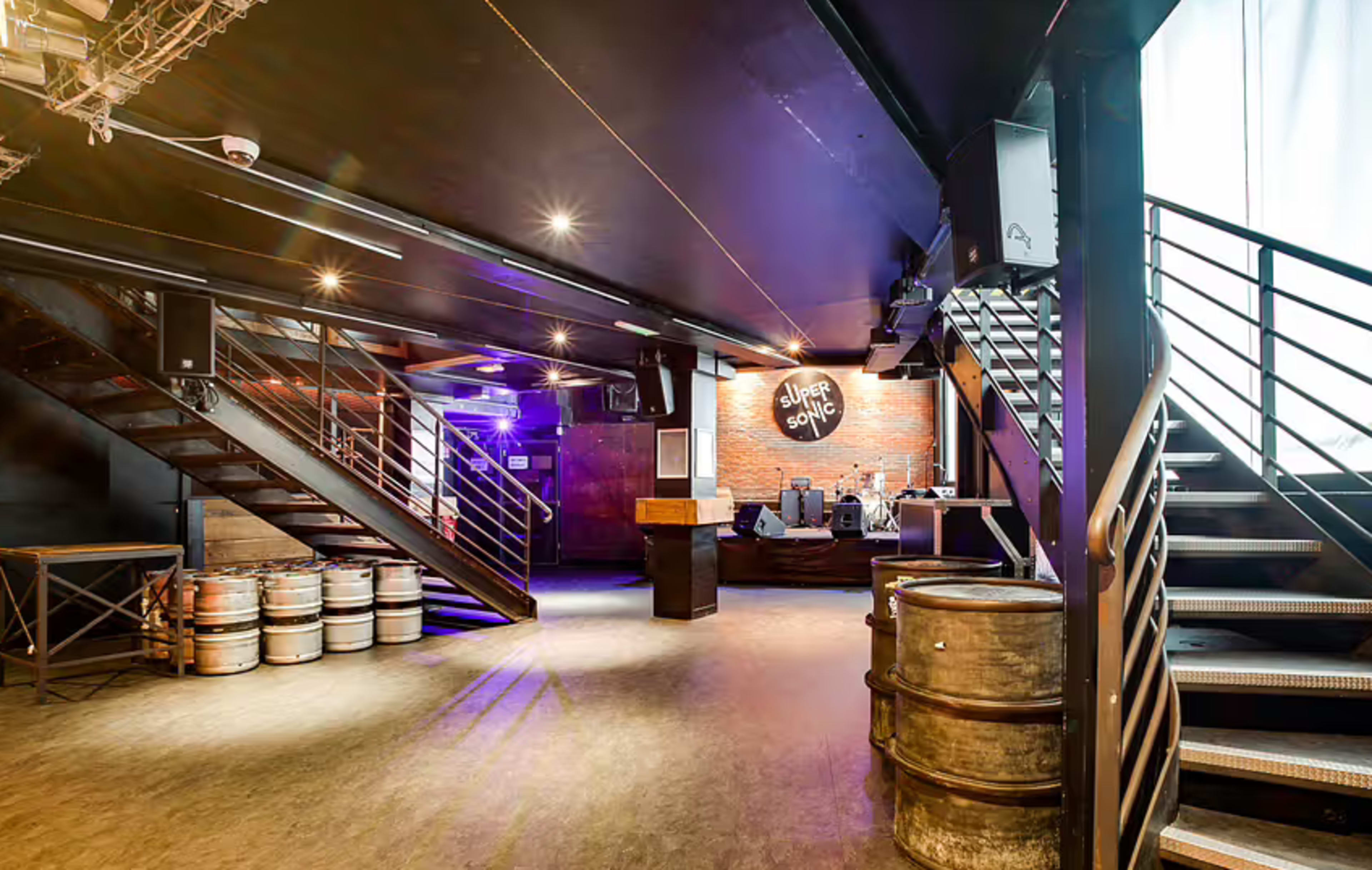 The image shows an interior of a music venue featuring two staircases, a stage with equipment in the background, and barrels positioned along the floor.