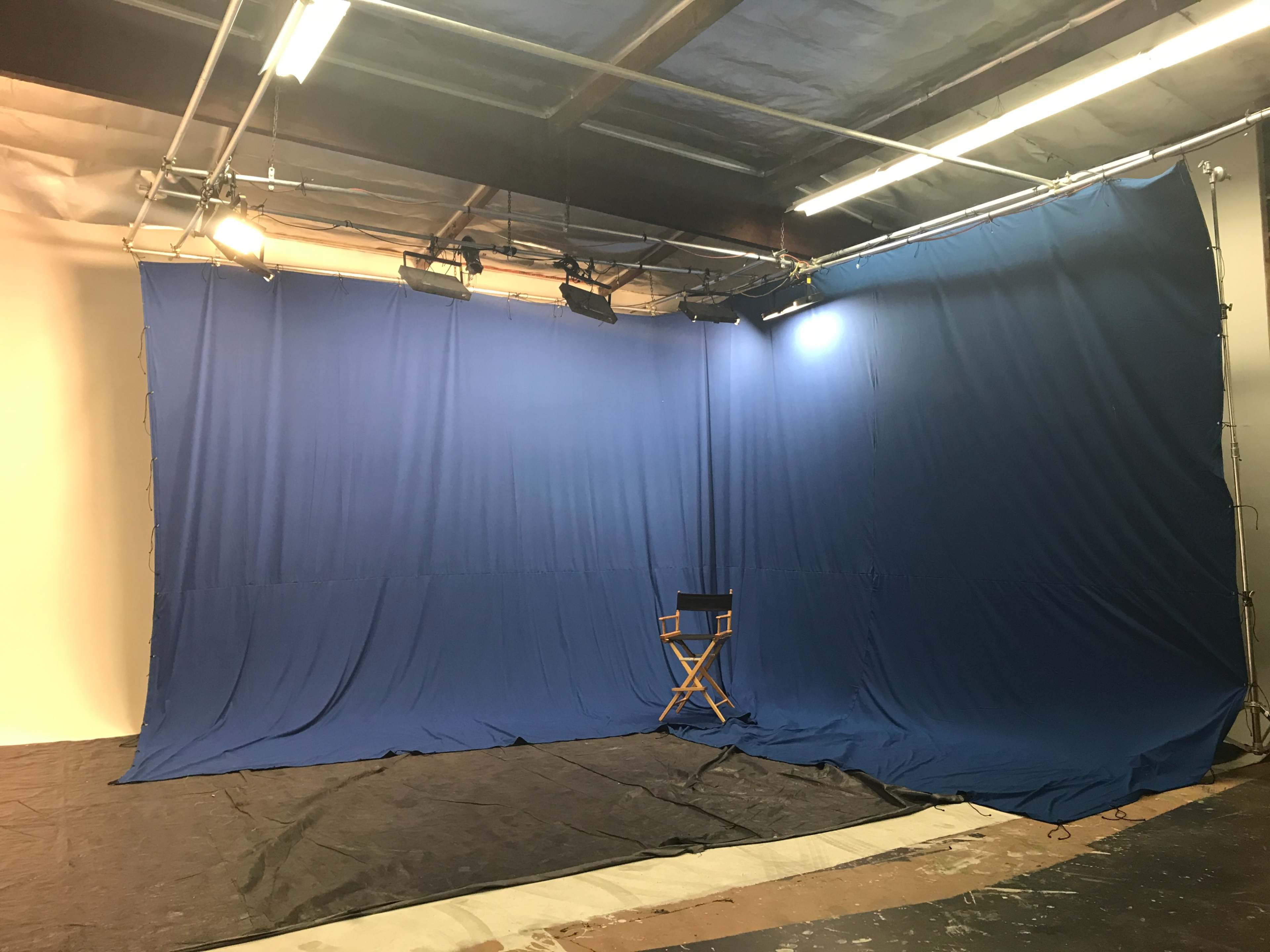 A photography studio featuring a large blue backdrop, lighting fixtures, and an empty director's chair on a black floor.