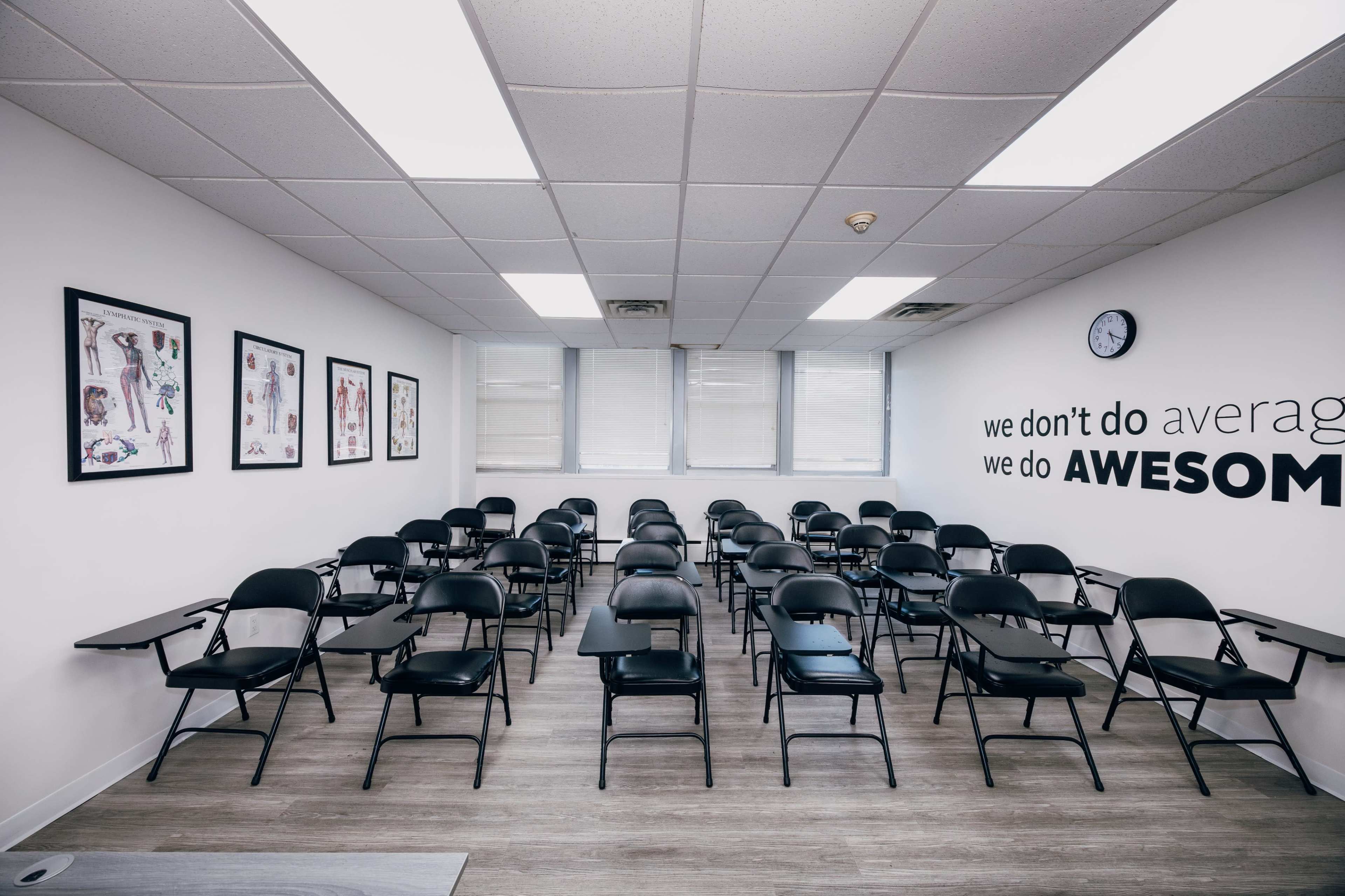 The image shows a classroom with rows of black chairs and desks, adorned with educational posters on the walls and a clock indicating the time.