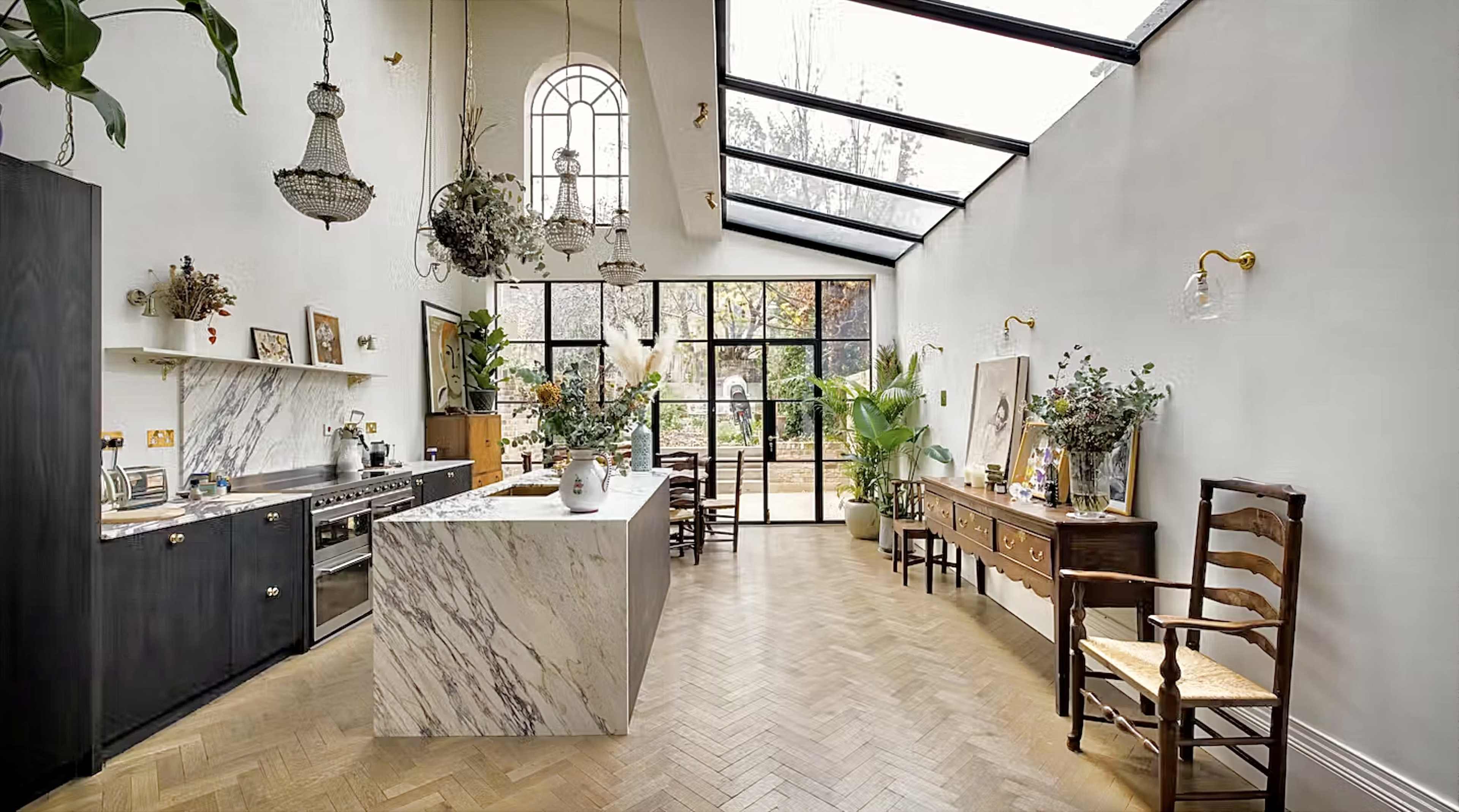 A spacious kitchen and dining area with a large marble island, high ceilings, and large glass doors leading to a garden.