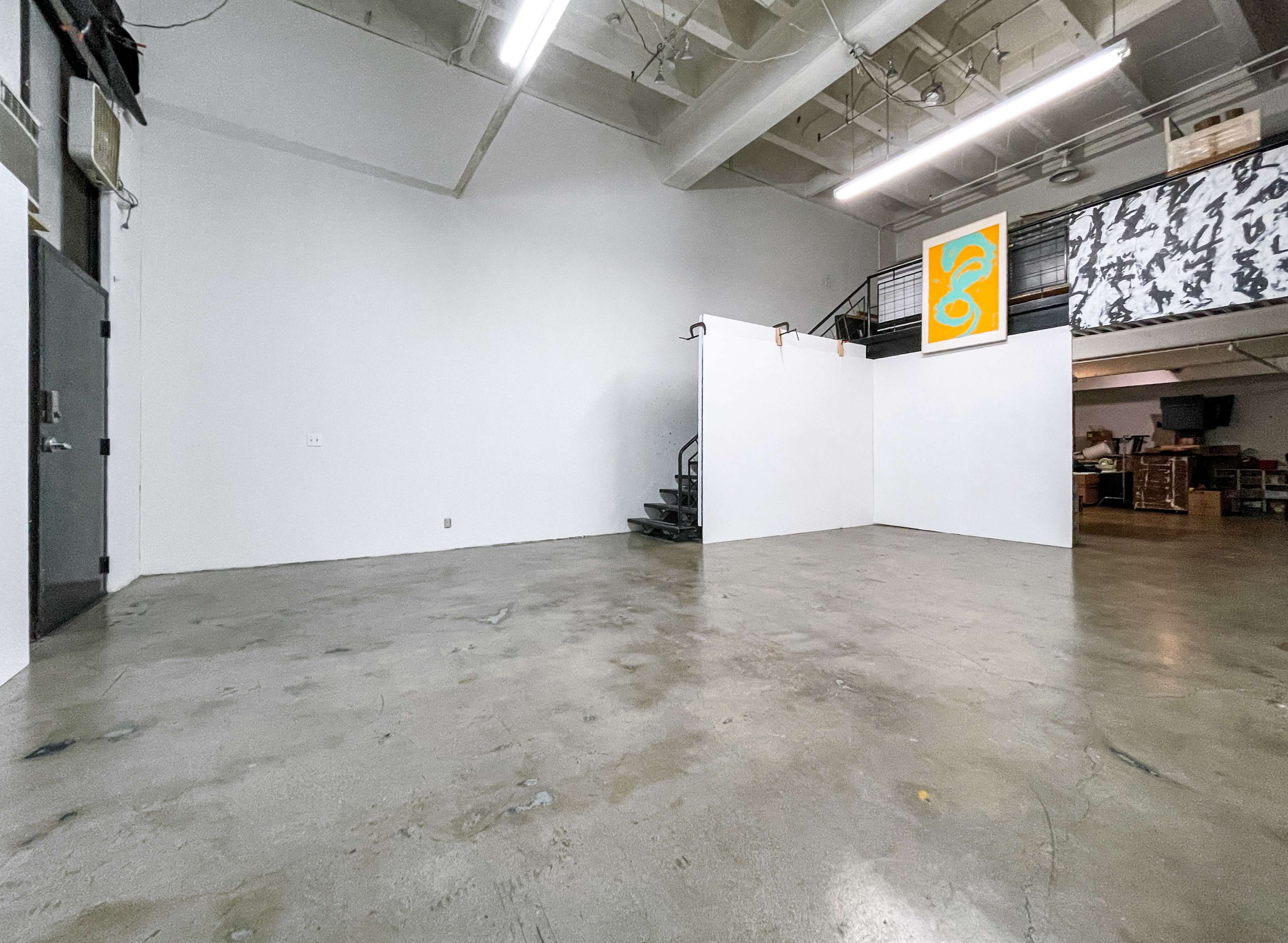 The image shows a spacious, empty room with a concrete floor, a staircase in the corner, and a large orange artwork mounted on the wall.