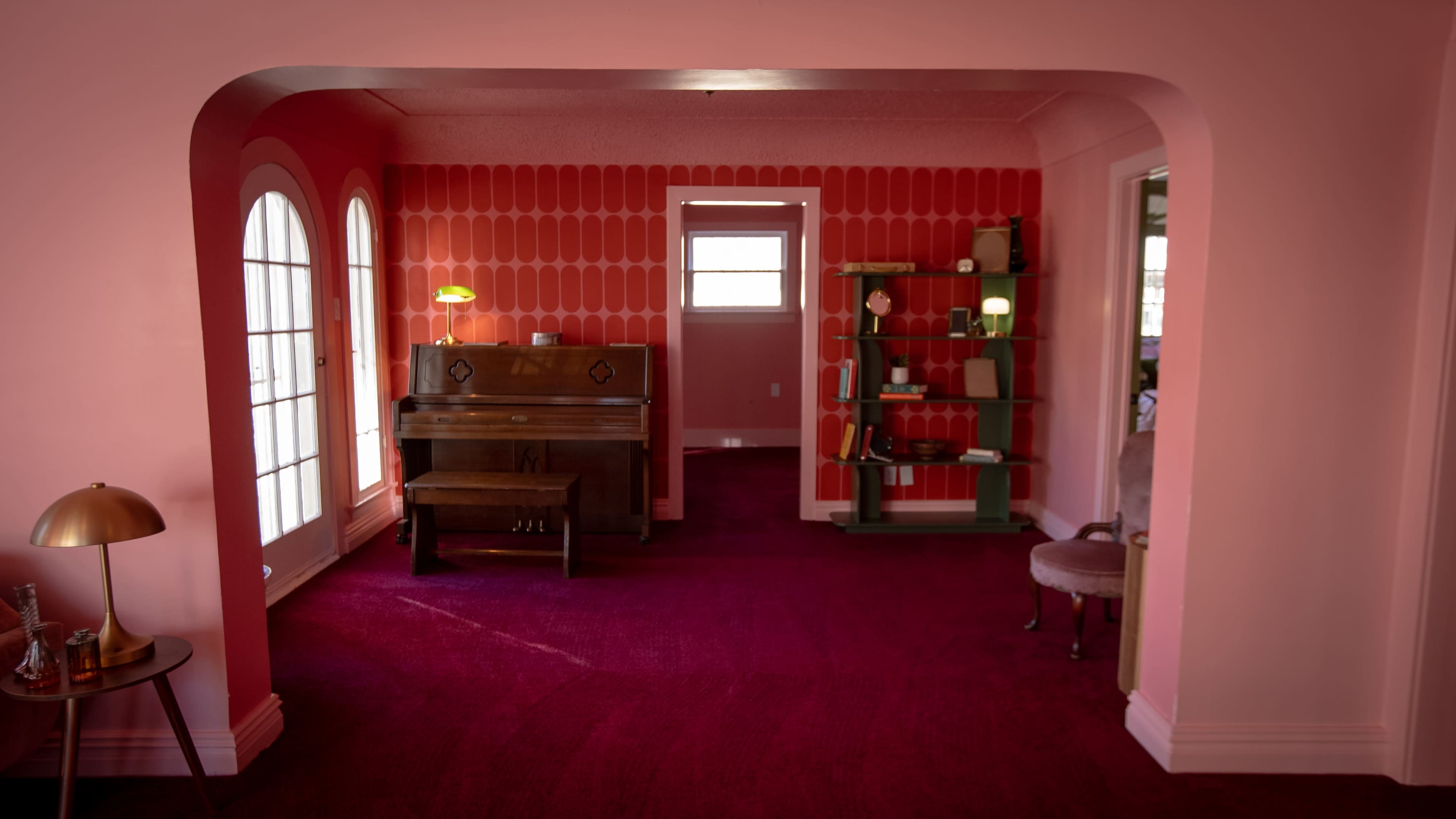 The image shows a bright pink room featuring a piano, a lamp, a bookshelf, and a plush carpet.