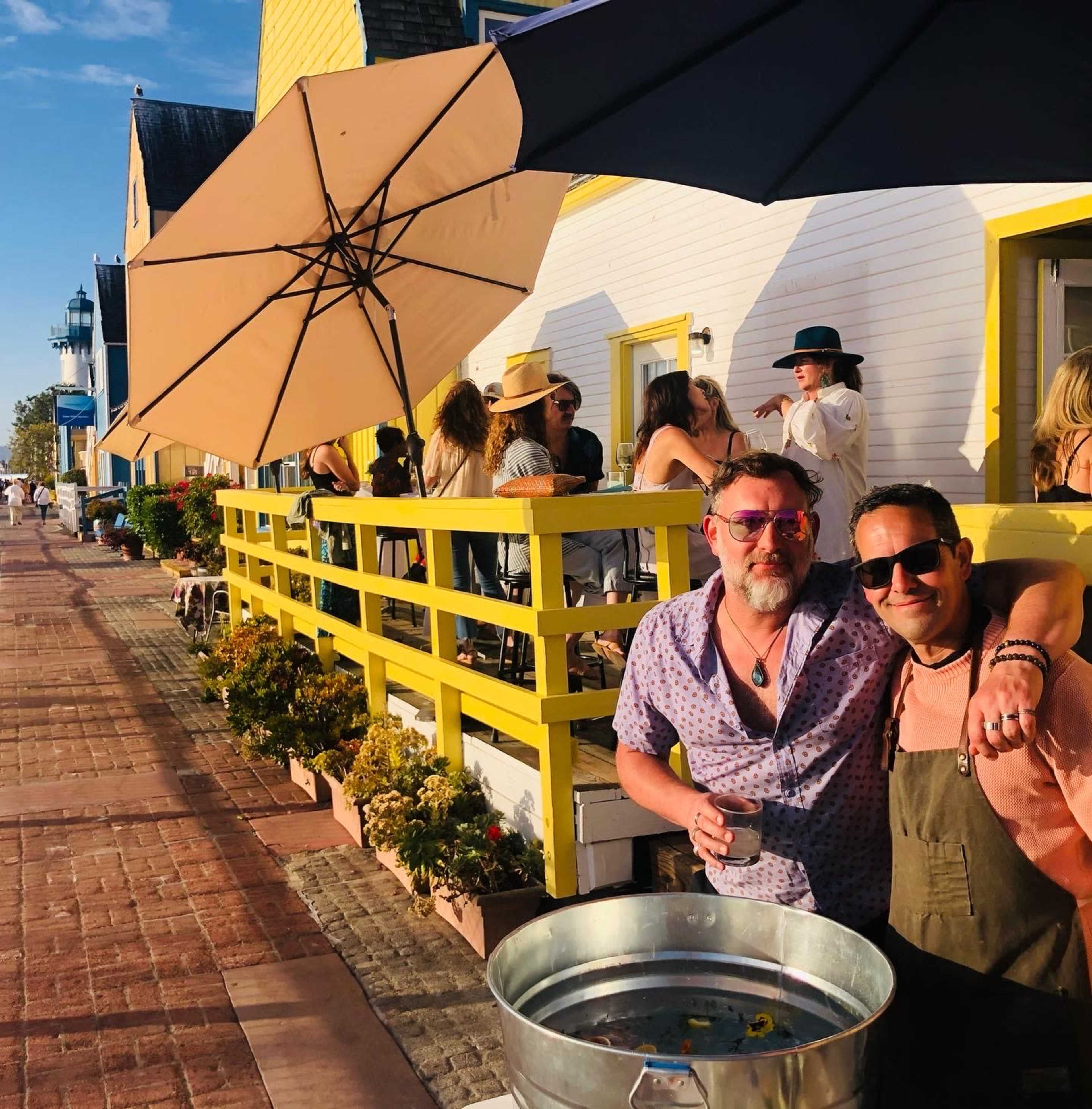 Two men pose in front of a vibrant yellow building with an outdoor gathering as people socialize on a sunny day.
