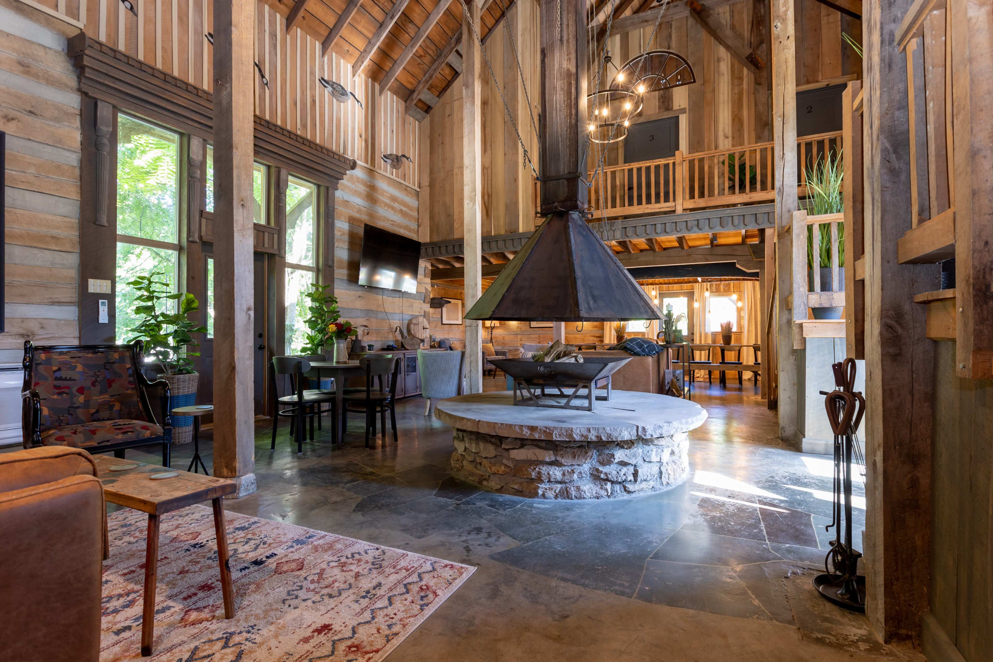 A spacious, rustic interior of a cabin with wooden walls, a large stone fireplace, and a central circular seating area.