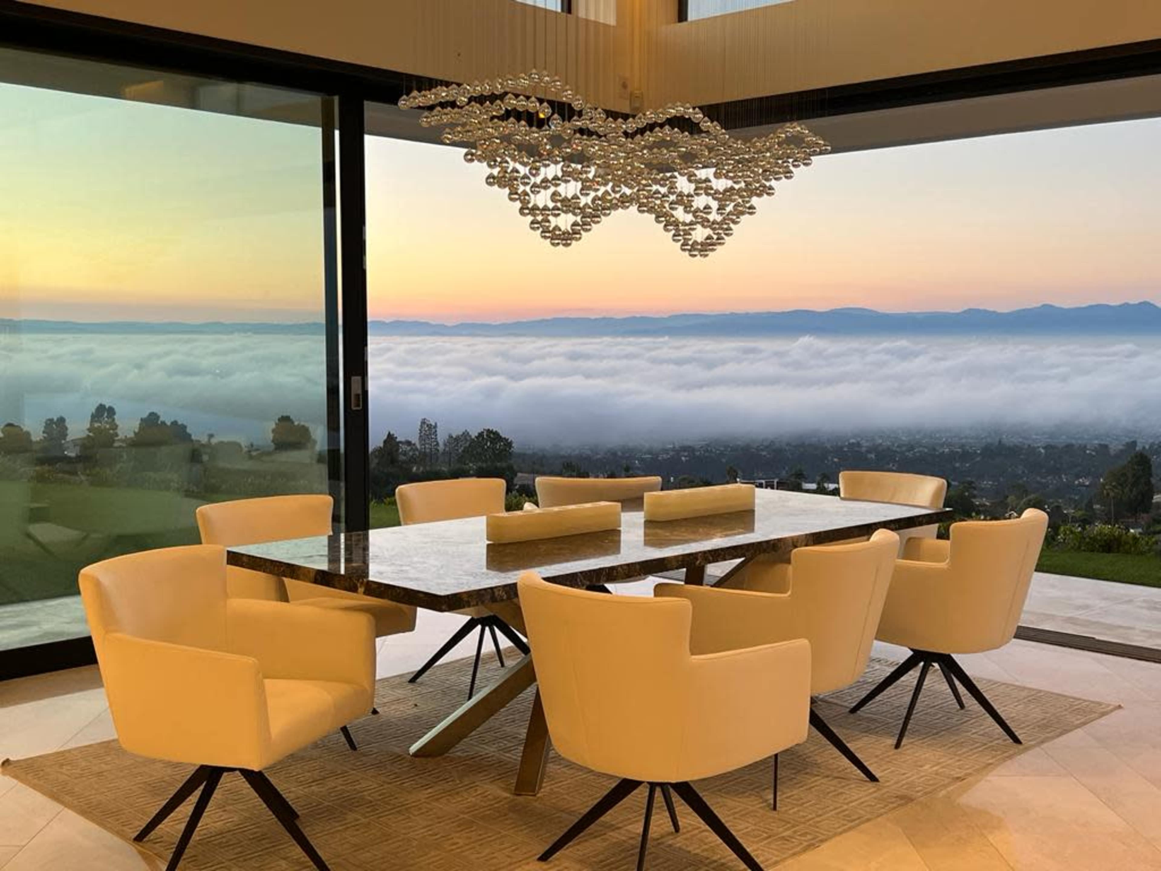 A modern dining room with a large table surrounded by eight chairs, overlooking a scenic view of clouds and mountains at sunset through large glass windows.