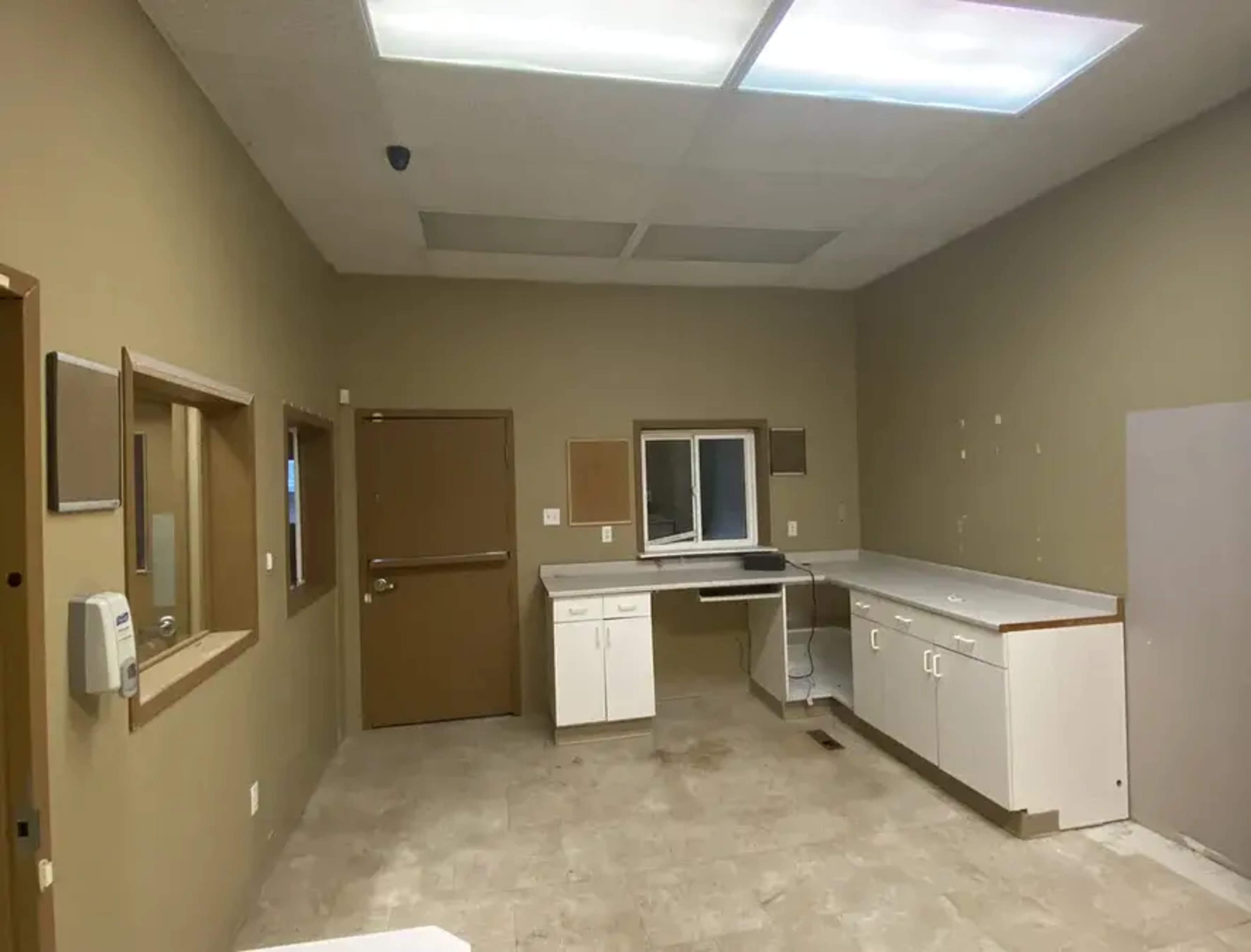 An empty, beige-walled room with a sink, countertops, and two windows, one of which is mounted in a wall.