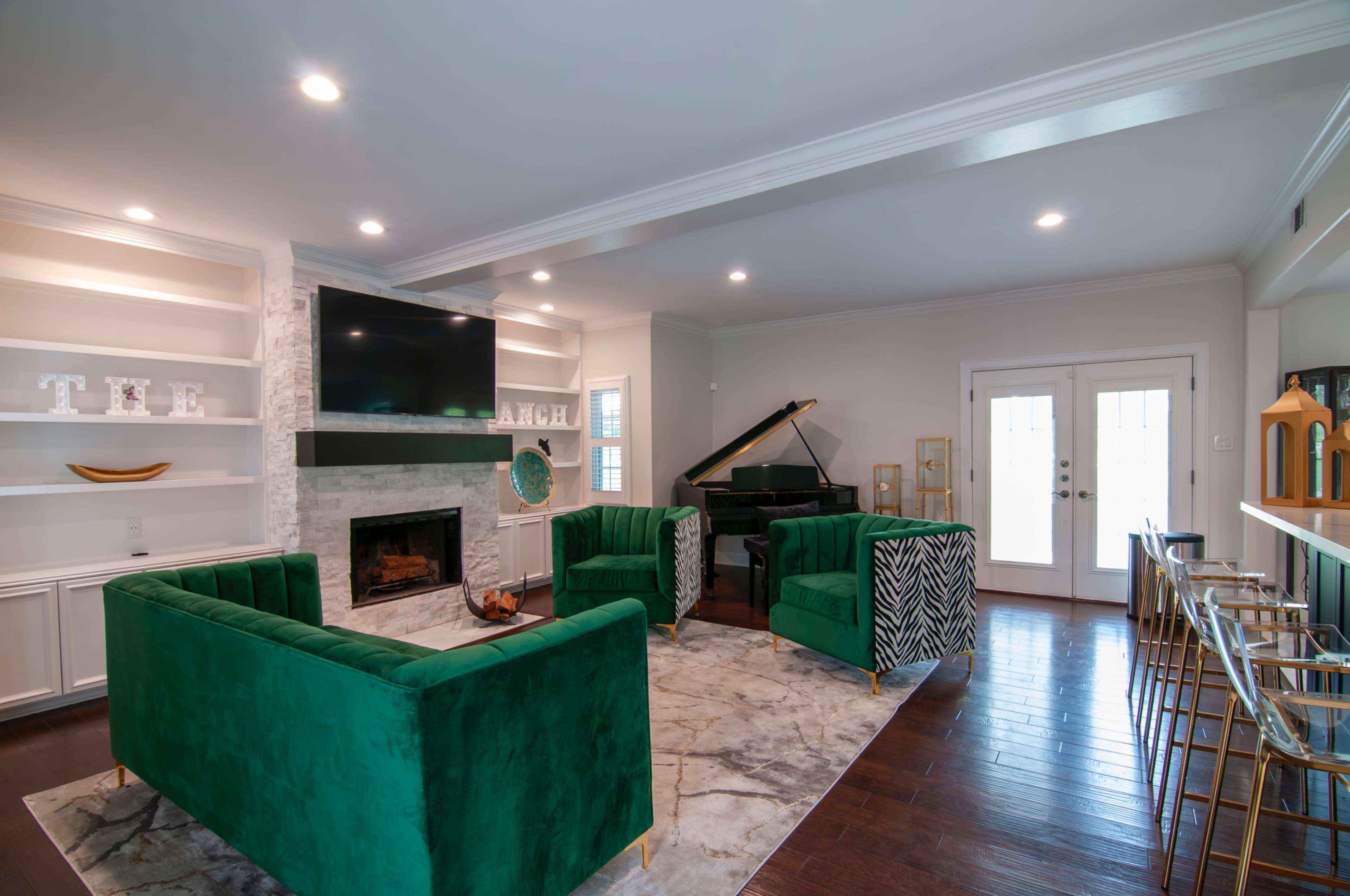 The image shows a modern living room with green upholstered chairs, a marble floor, a vertical black piano, a stone fireplace, and a wall-mounted television.