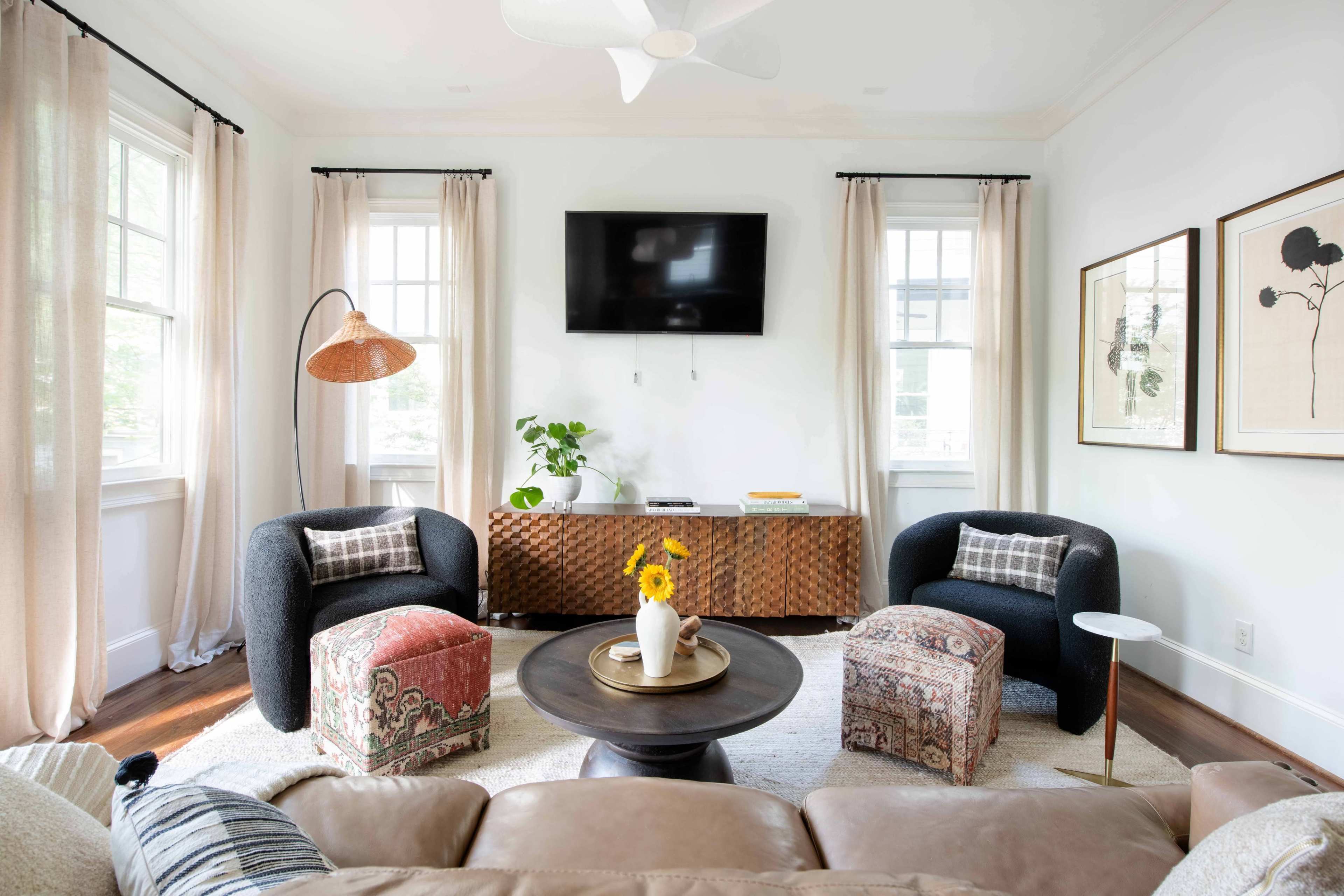 The living room features two dark armchairs, a round coffee table, and a wooden console under a wall-mounted TV, with natural light coming through the large windows.