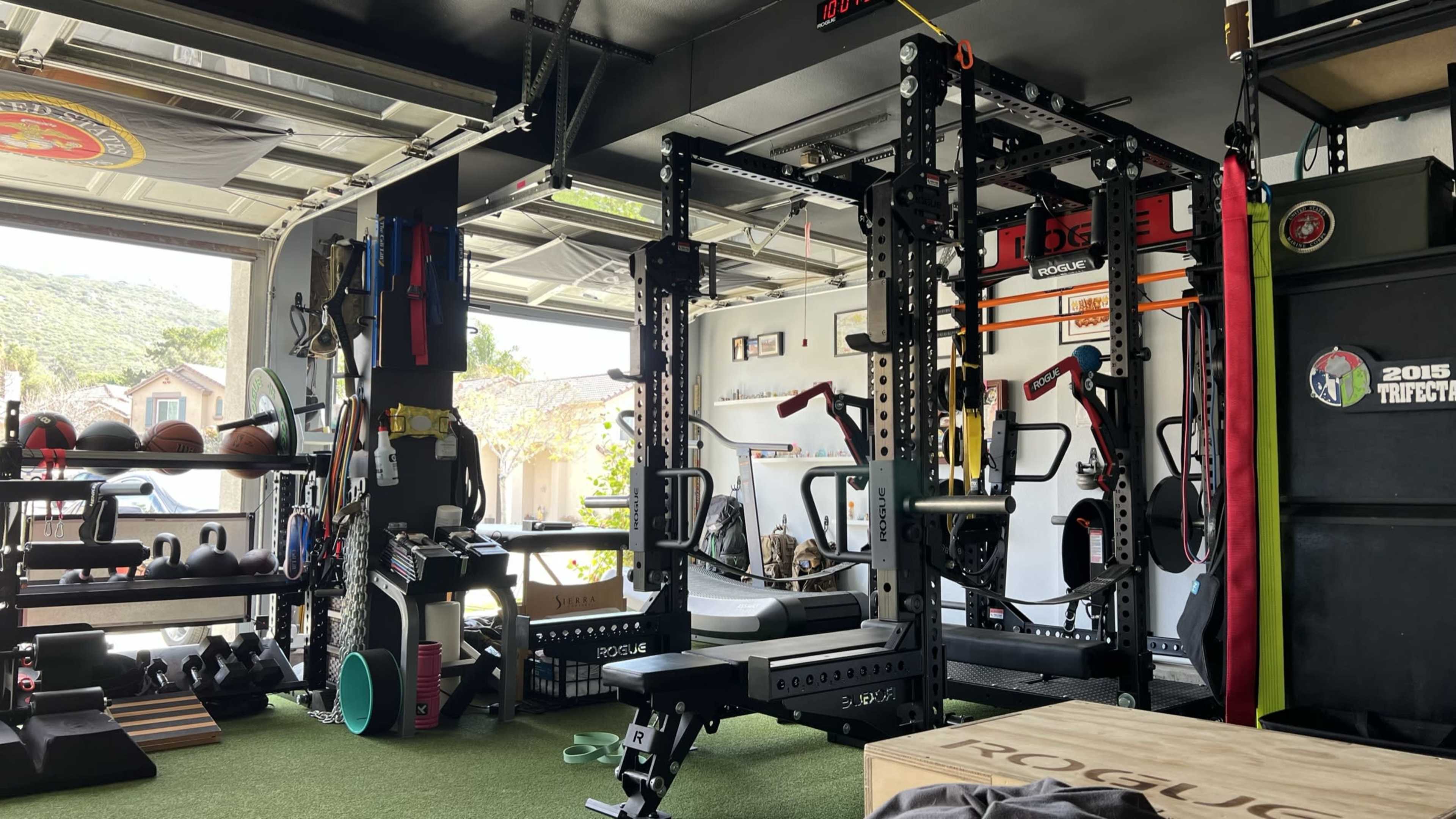 The image shows a well-equipped home gym in a garage, featuring various exercise machines, weights, and exercise accessories arranged neatly.