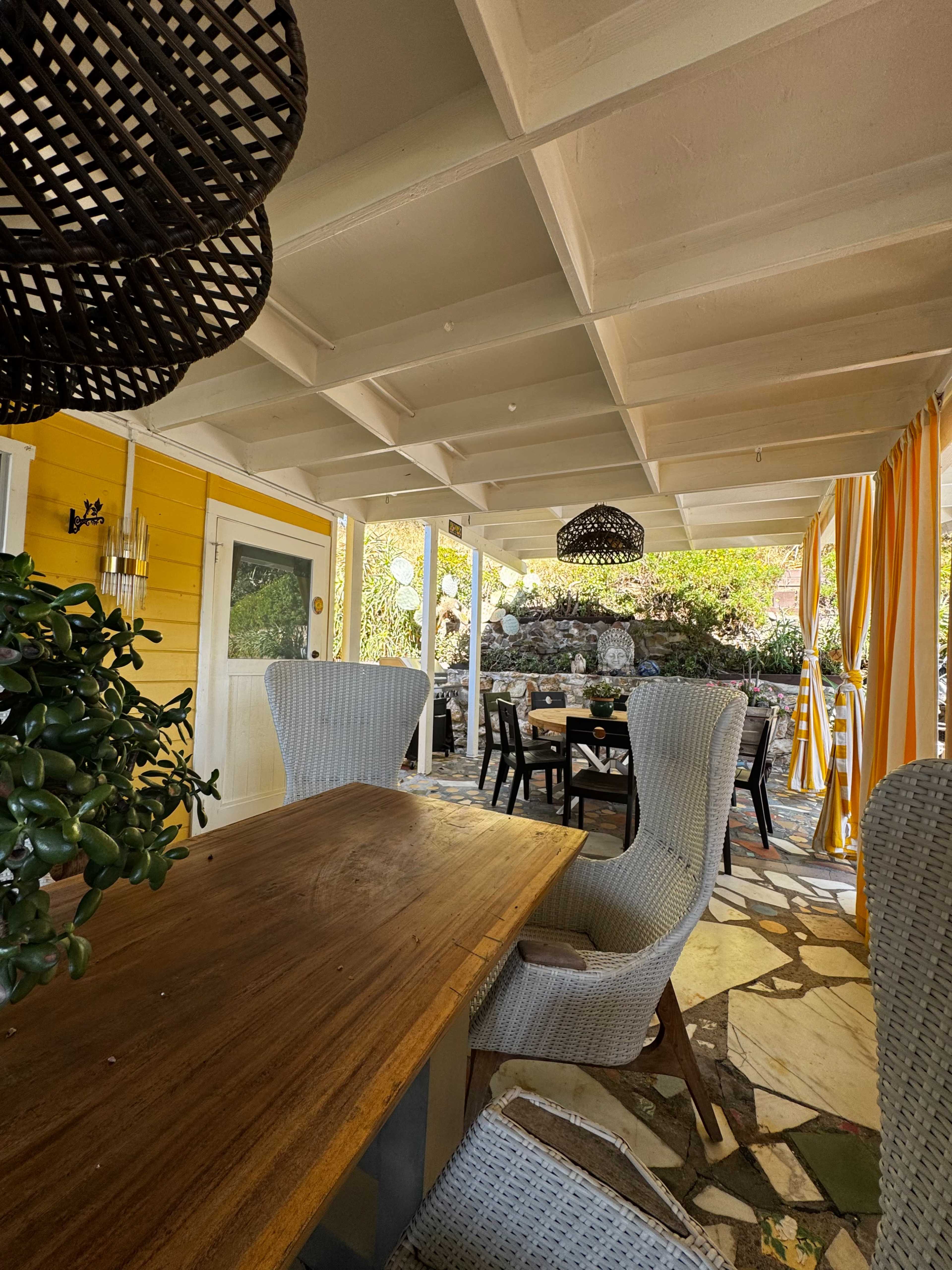 The image shows a patio area with a wooden table and wicker chairs, surrounded by colorful drapes and a stone floor.
