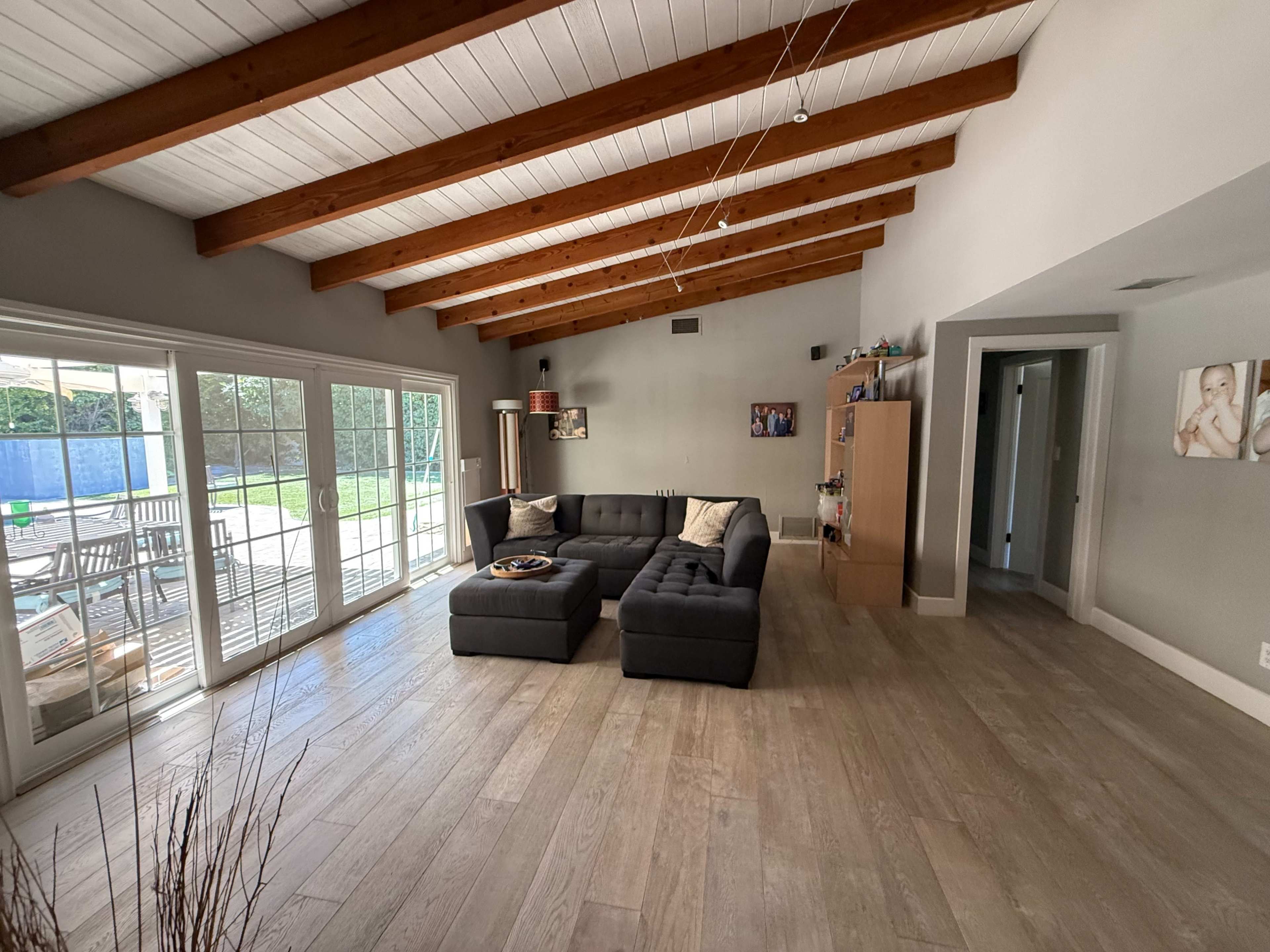 A spacious living room features a gray sectional sofa, large windows opening to a backyard, and wooden beams on the ceiling.