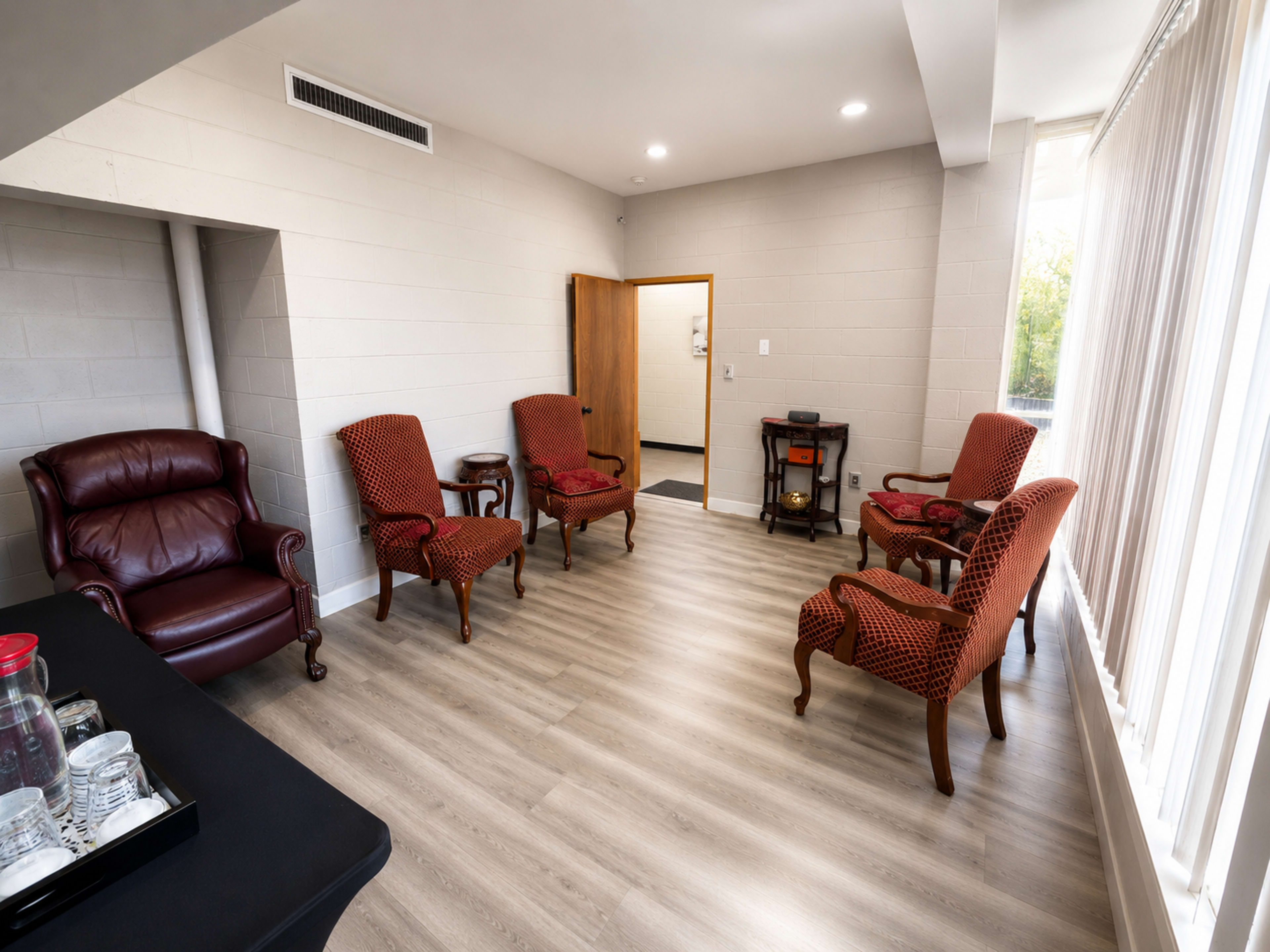 The image shows a waiting room furnished with several armchairs, a wooden side table, and a small fireplace, all situated in a well-lit space with large windows.
