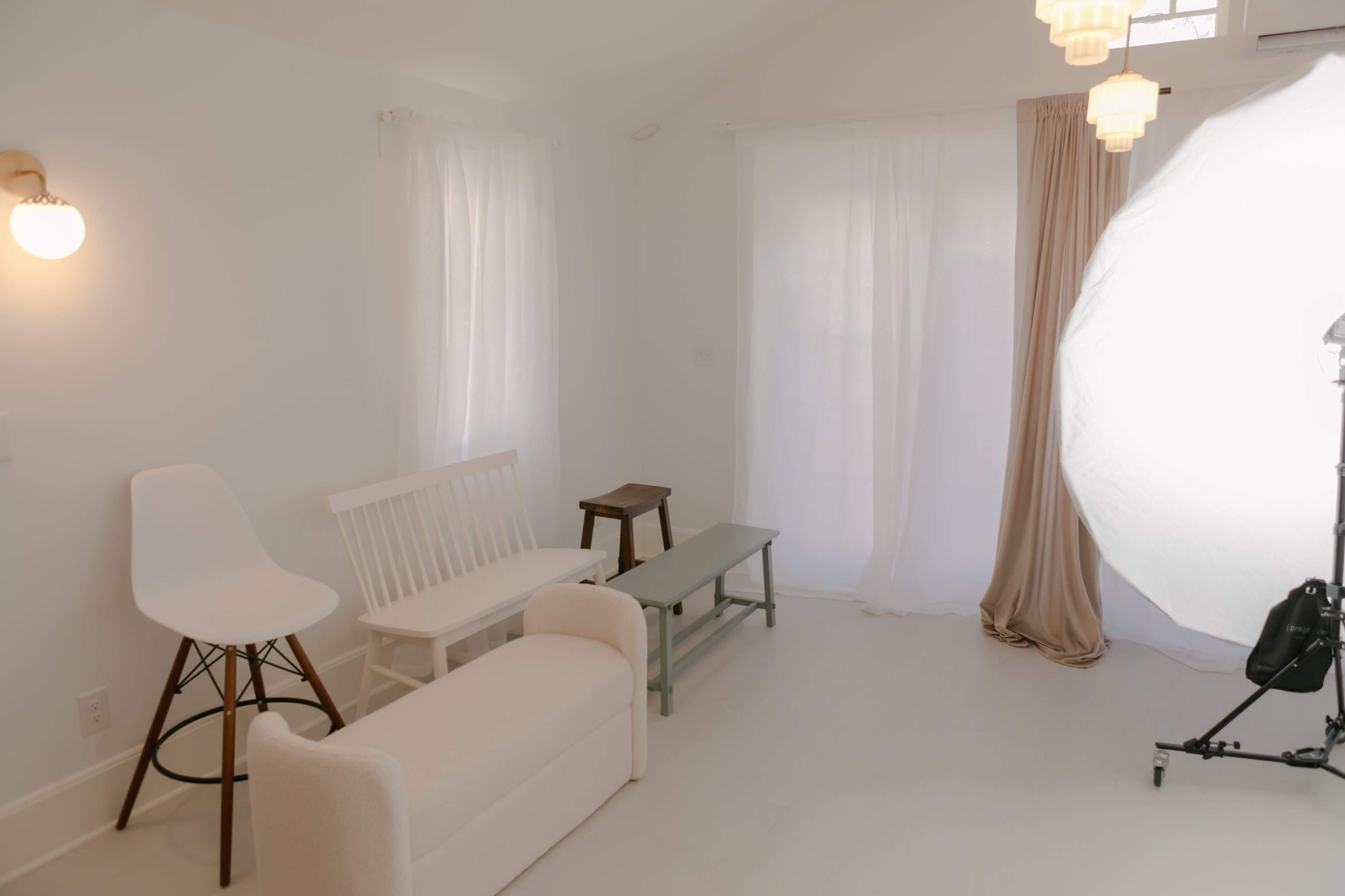 The image shows a minimalist interior space with a white color scheme, featuring a white sofa, a wooden bench, a green coffee table, and a light source from a large diffused lamp.