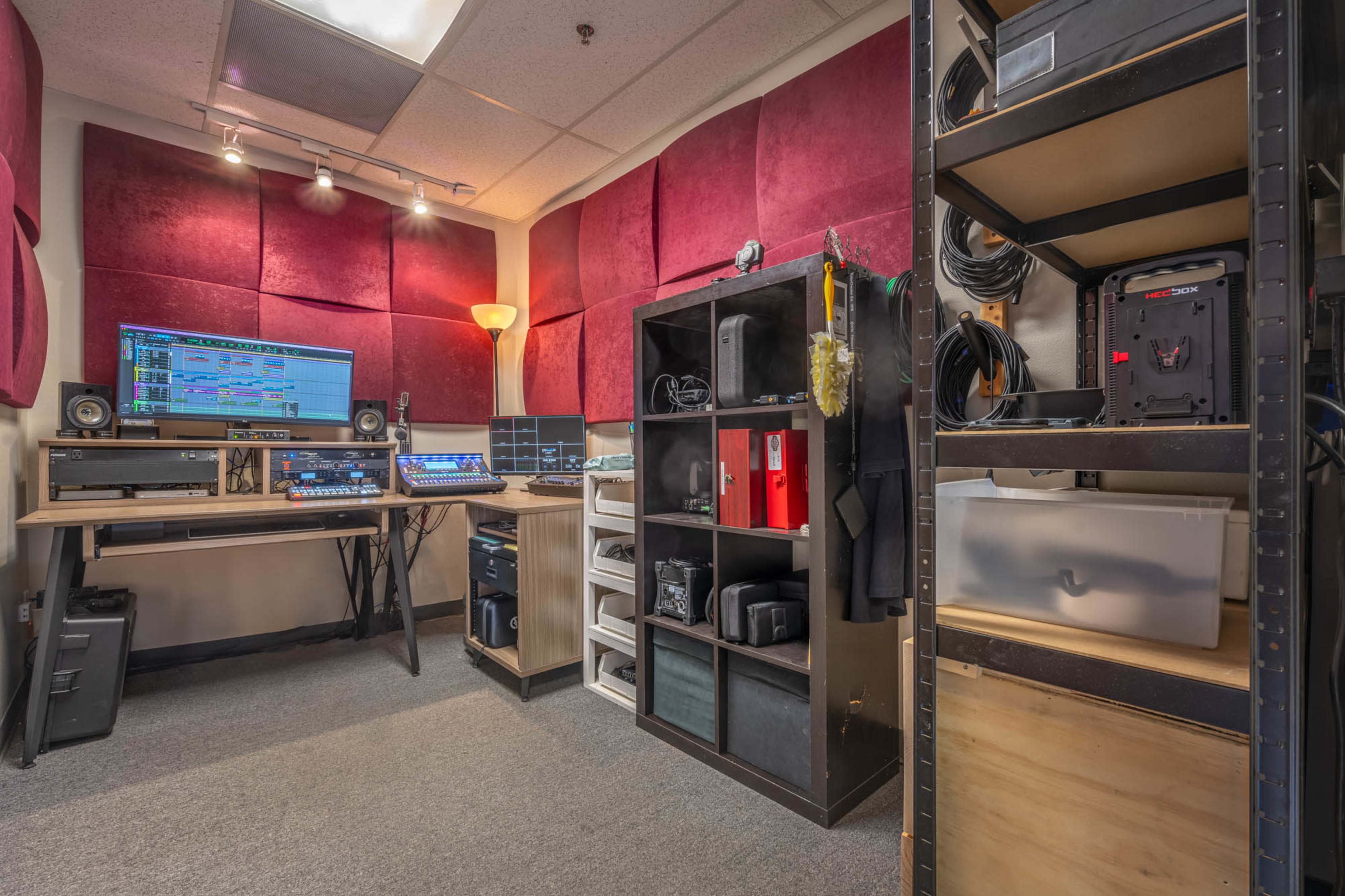 A small studio room features equipment for music production, including a computer setup, shelves with gear, and soundproofing panels on the walls.