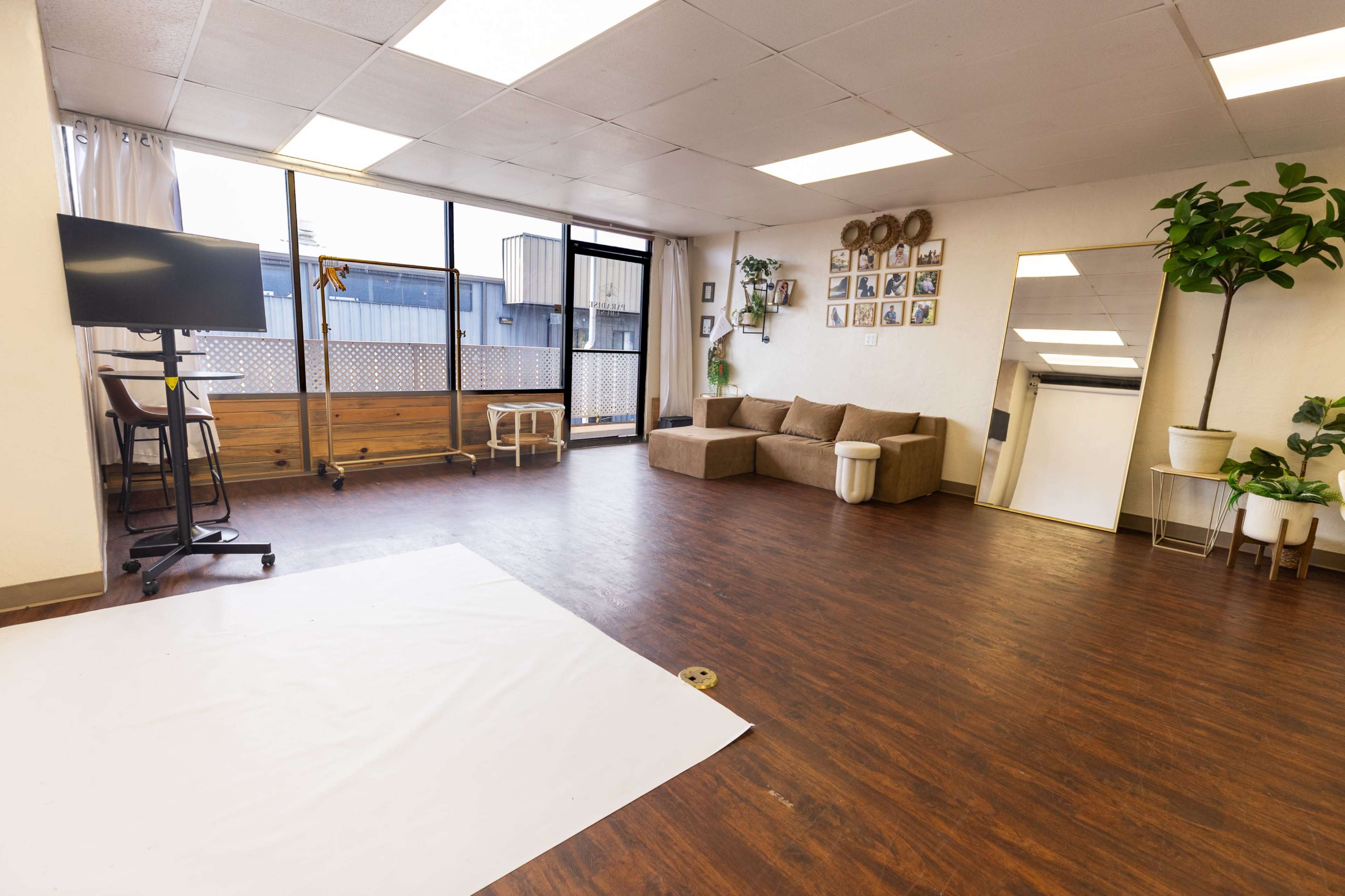 The image shows a spacious, well-lit interior with wooden flooring, a large window, a sectional couch, a television on a stand, and several framed photos on the wall.