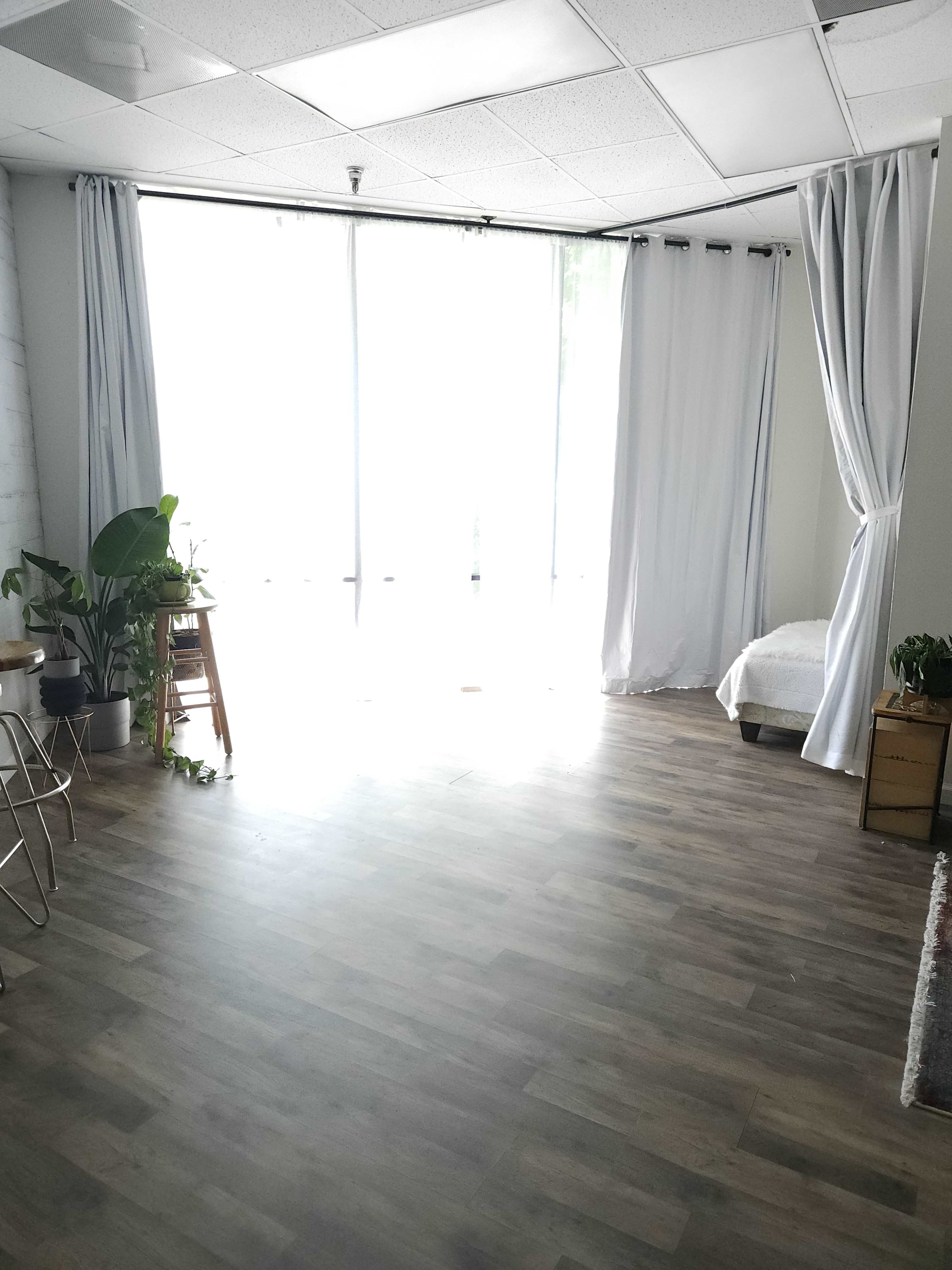 A bright room with large windows, light-colored curtains, wooden flooring, and minimal furnishings including plants and a bed.