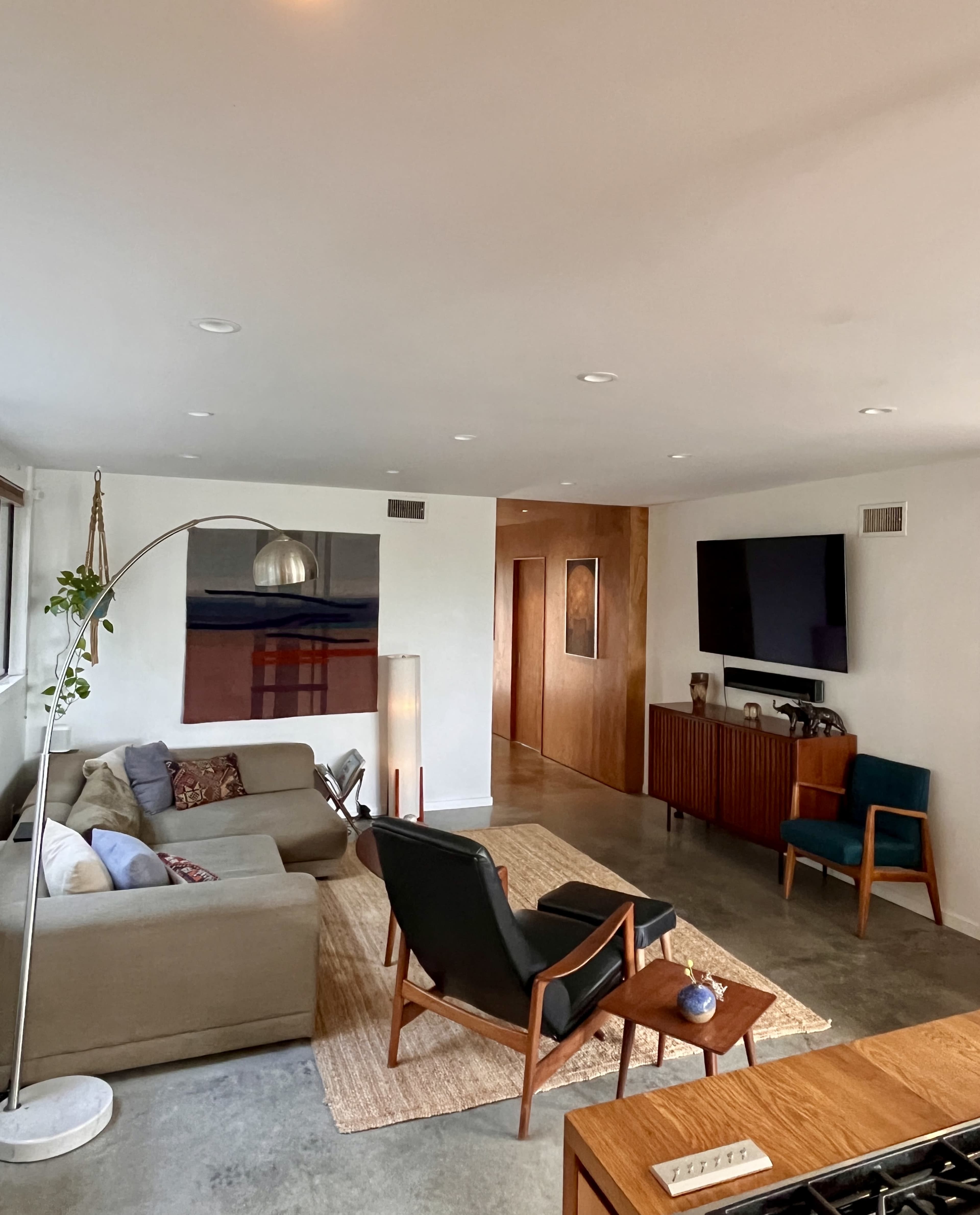 The image shows a modern living room with a sofa, two chairs, a floor lamp, and a TV mounted on the wall, alongside a wooden console and a woven rug on a concrete floor.