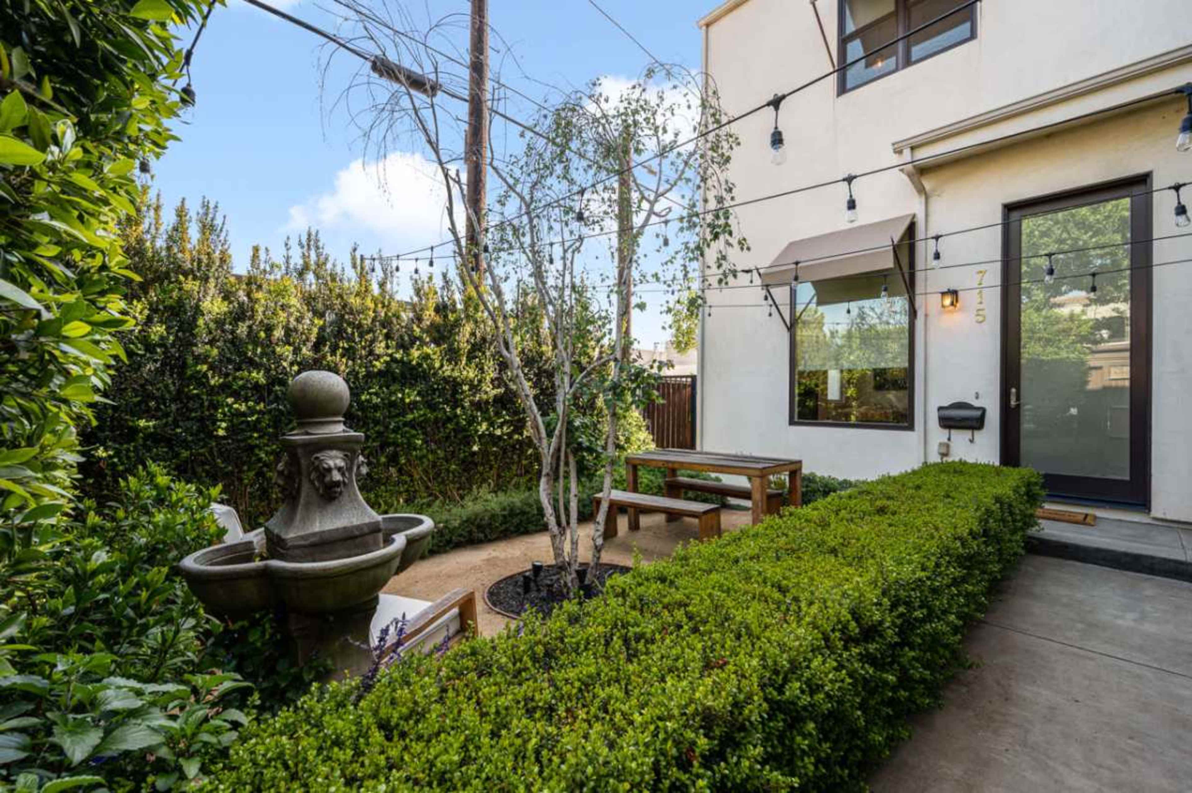 The image shows a small courtyard with a decorative fountain, bordered by neatly trimmed hedges and outdoor seating.