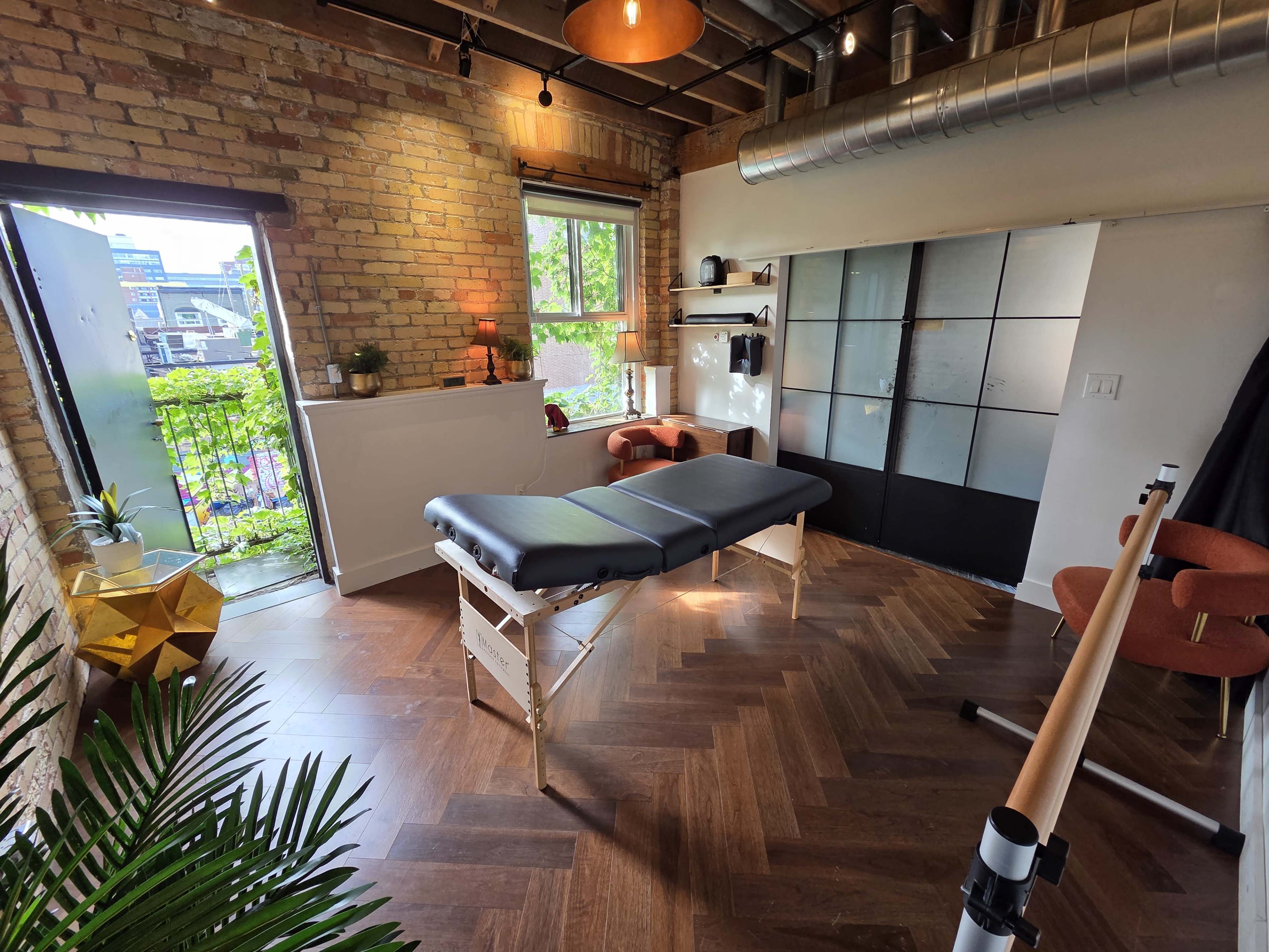 The image depicts a massage therapy room featuring a massage table, wooden flooring, exposed brick walls, and a window that opens to a small outdoor area.