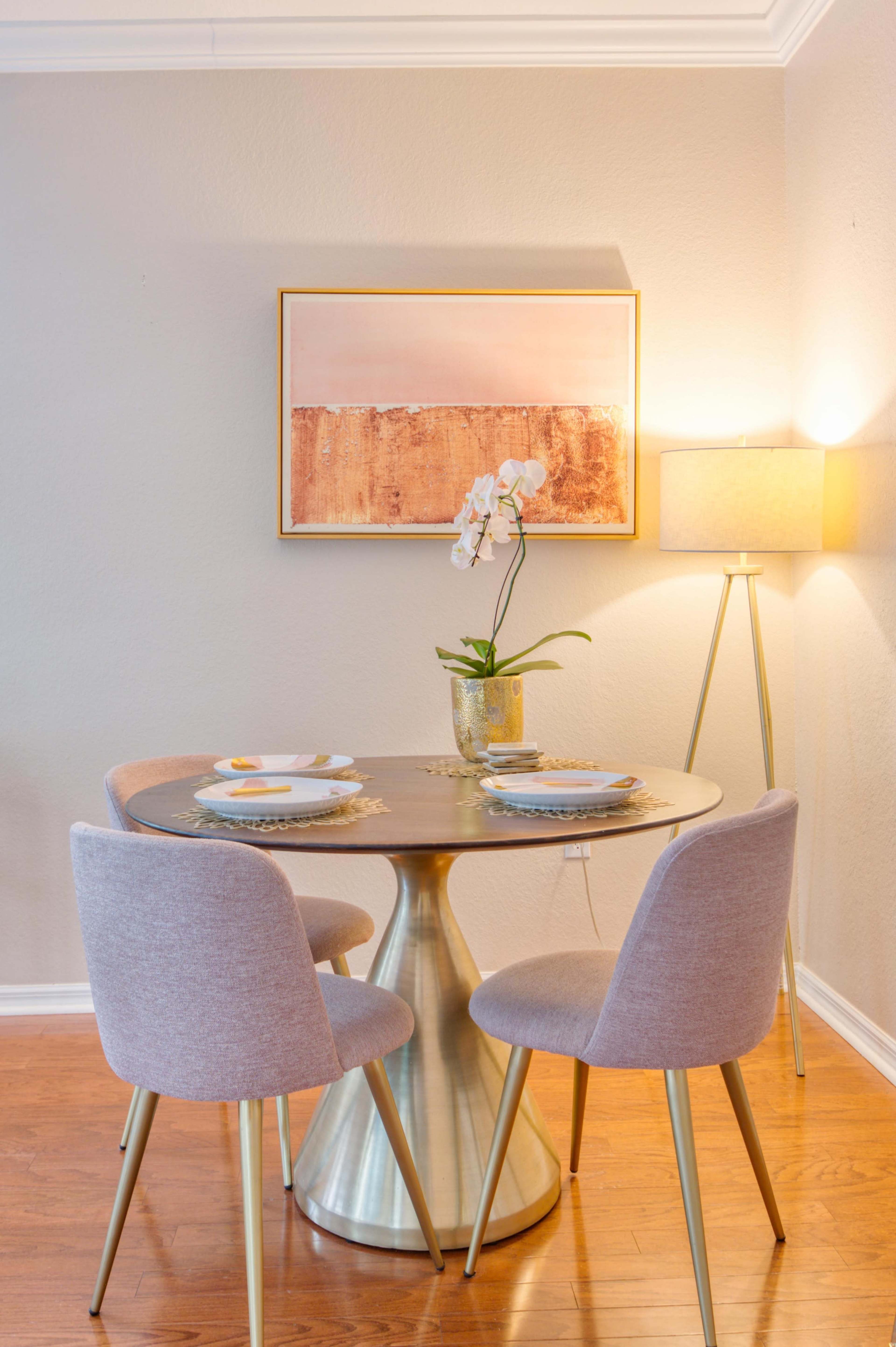 A round dining table with two chairs, an orchid in a vase, and a lamp stands in a well-lit corner with a framed artwork on the wall.