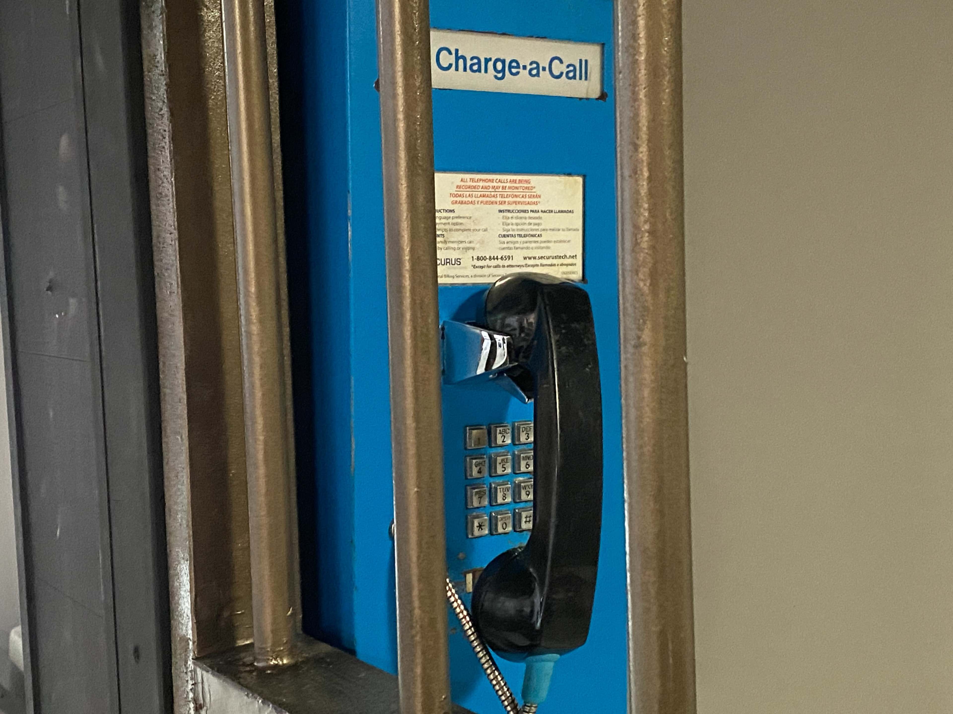 The image shows a payphone enclosed in metal bars with a bright blue panel labeled "Charge-a-Call."