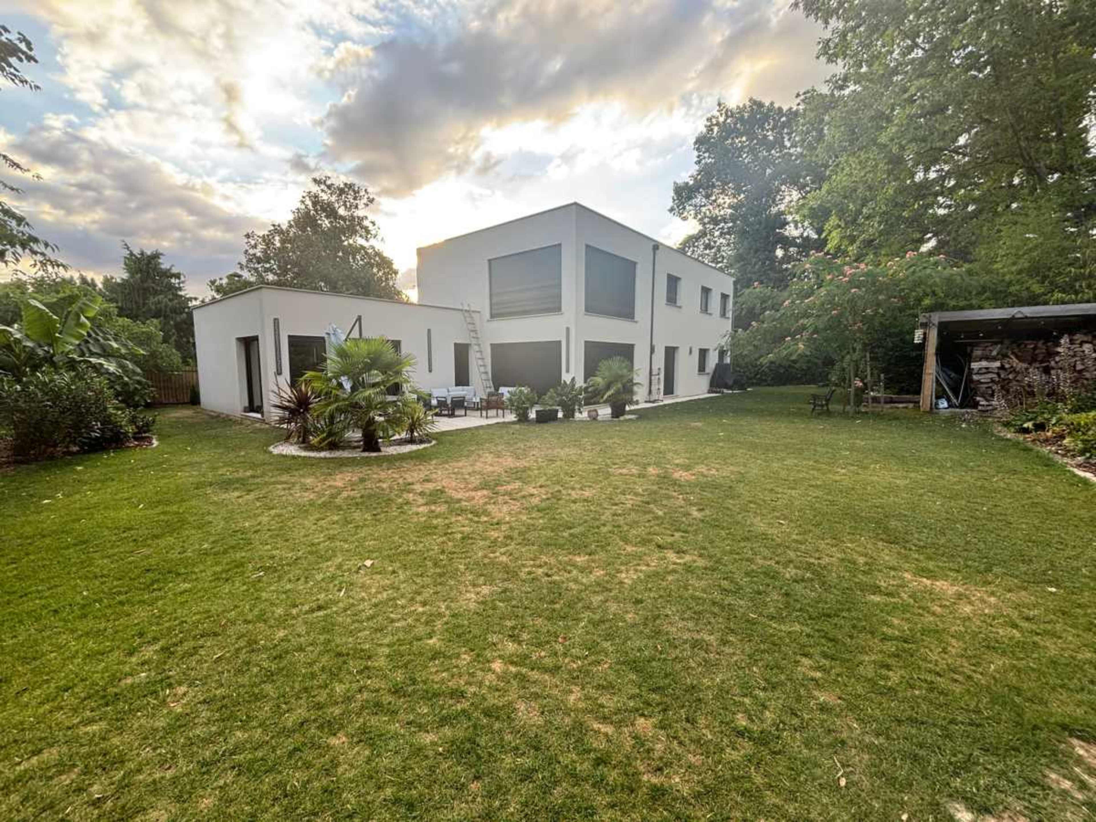 The image shows a modern two-story house surrounded by a grassy yard, with a patio area and various plants.